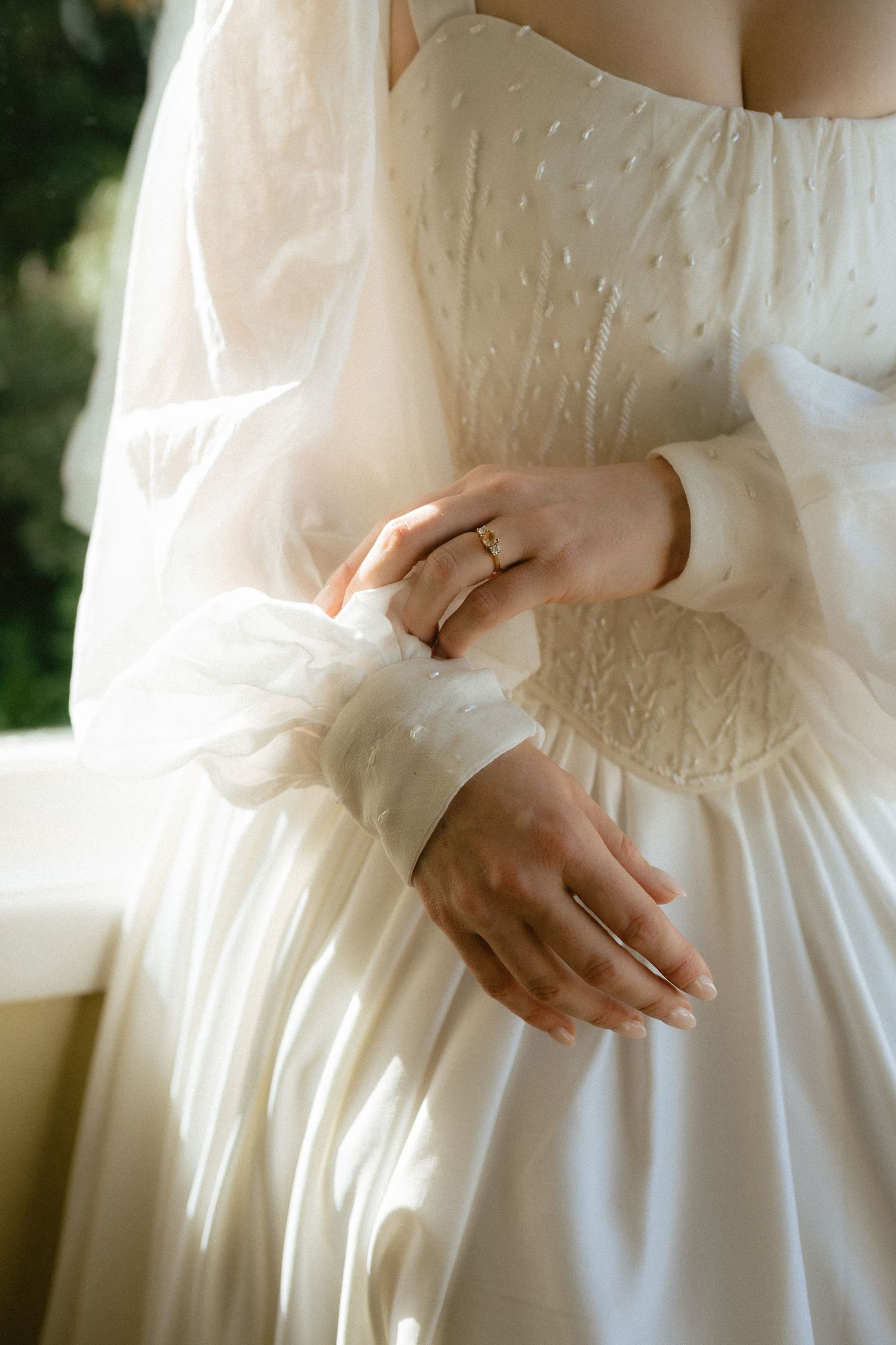 Bride in elegant white wedding dress with sheer sleeves adjusting her gold ring on her finger.