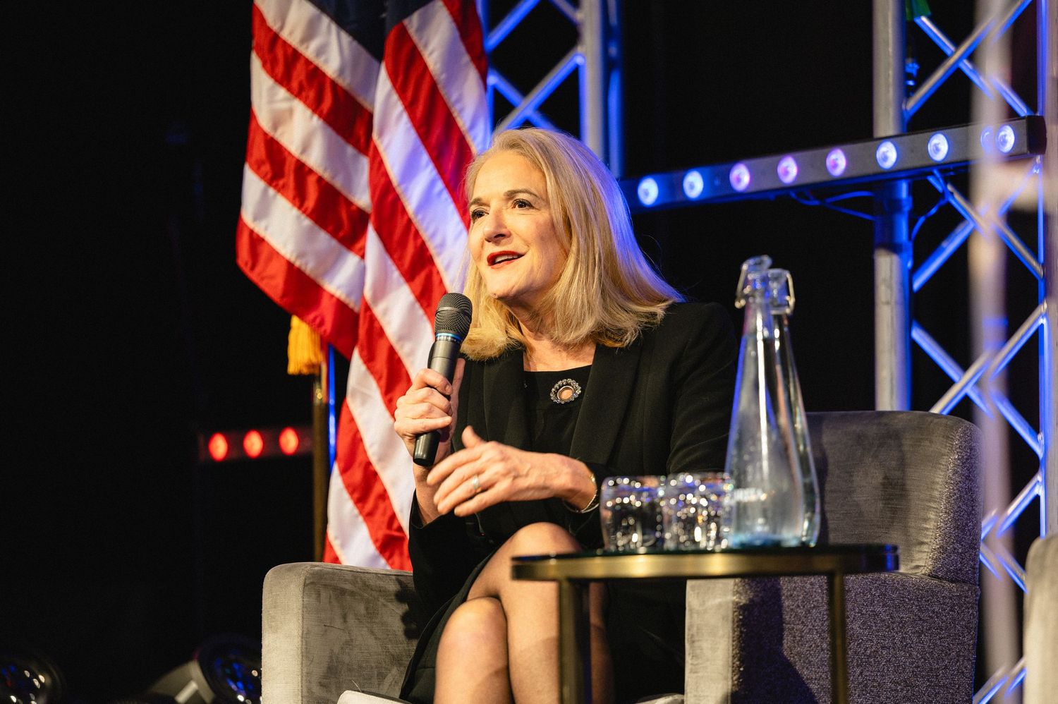 Speaker stands at podium with American flag backdrop during political event with stage lighting.