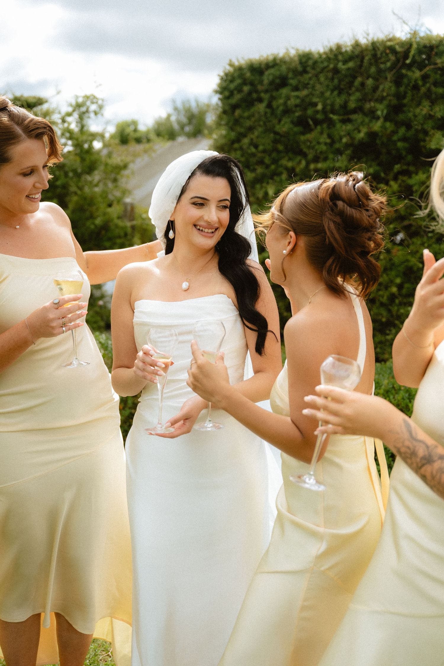 Bride in white dress and veil celebrates with bridesmaids in yellow dresses, holding champagne glasses outdoors.