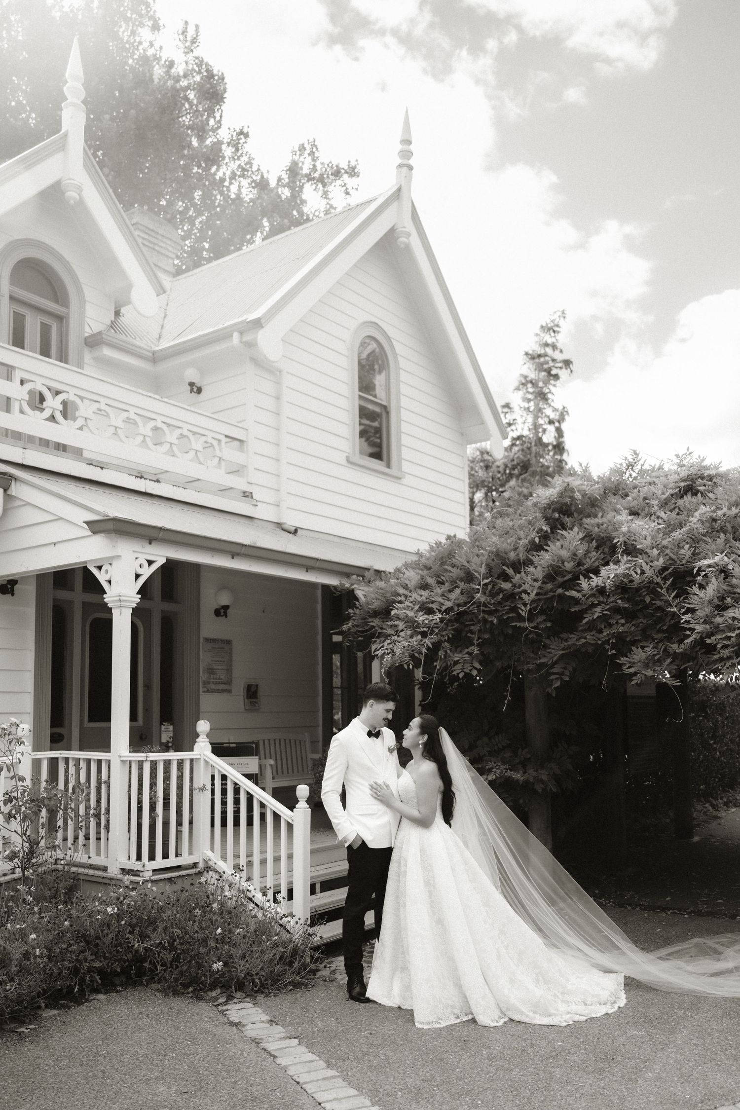 Black and white photo of a bride and groom posing in front of a charming Victorian house with a flowing veil.