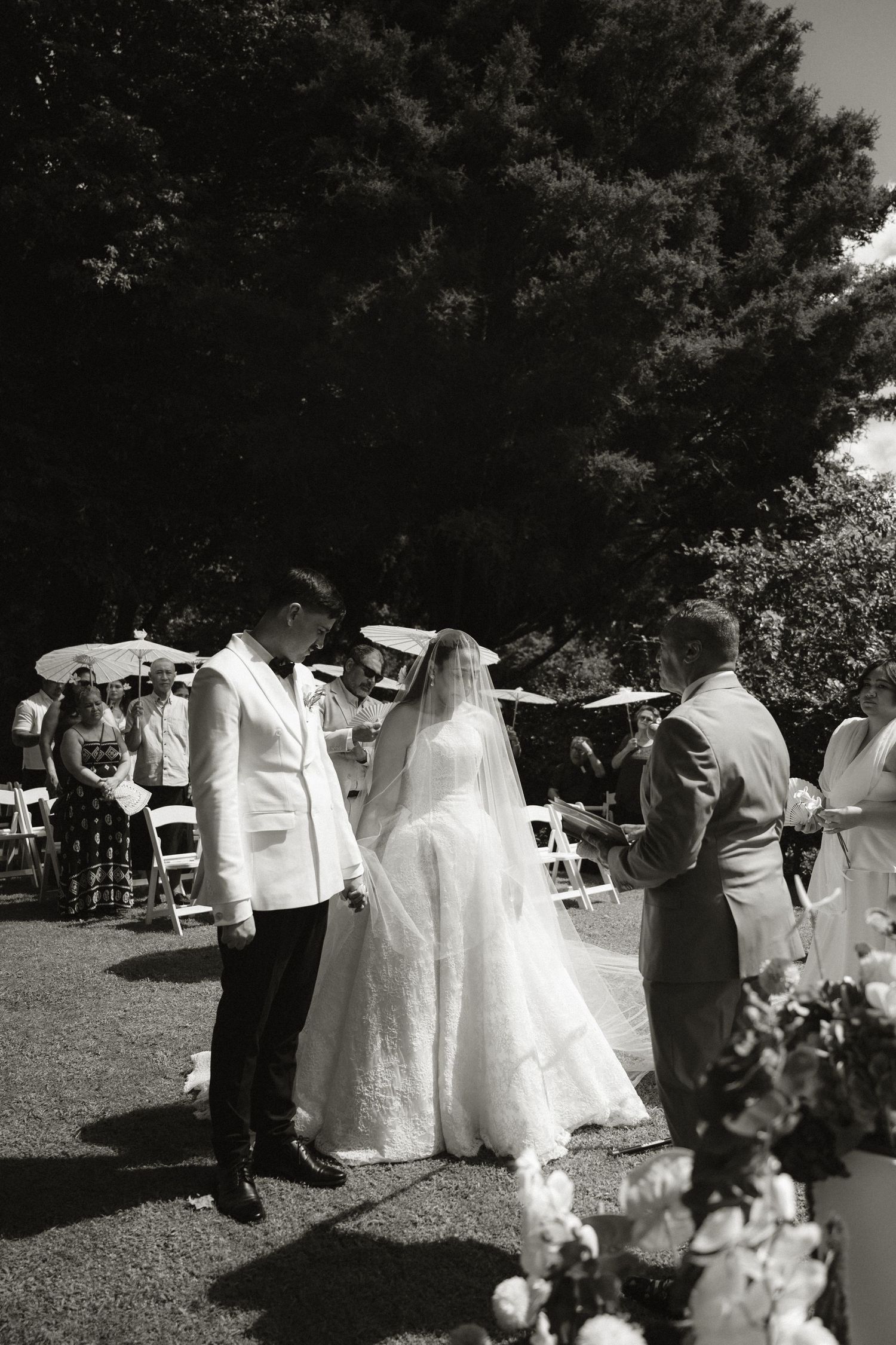 Black and white outdoor wedding ceremony with bride in flowing gown and groom in white suit exchanging vows.