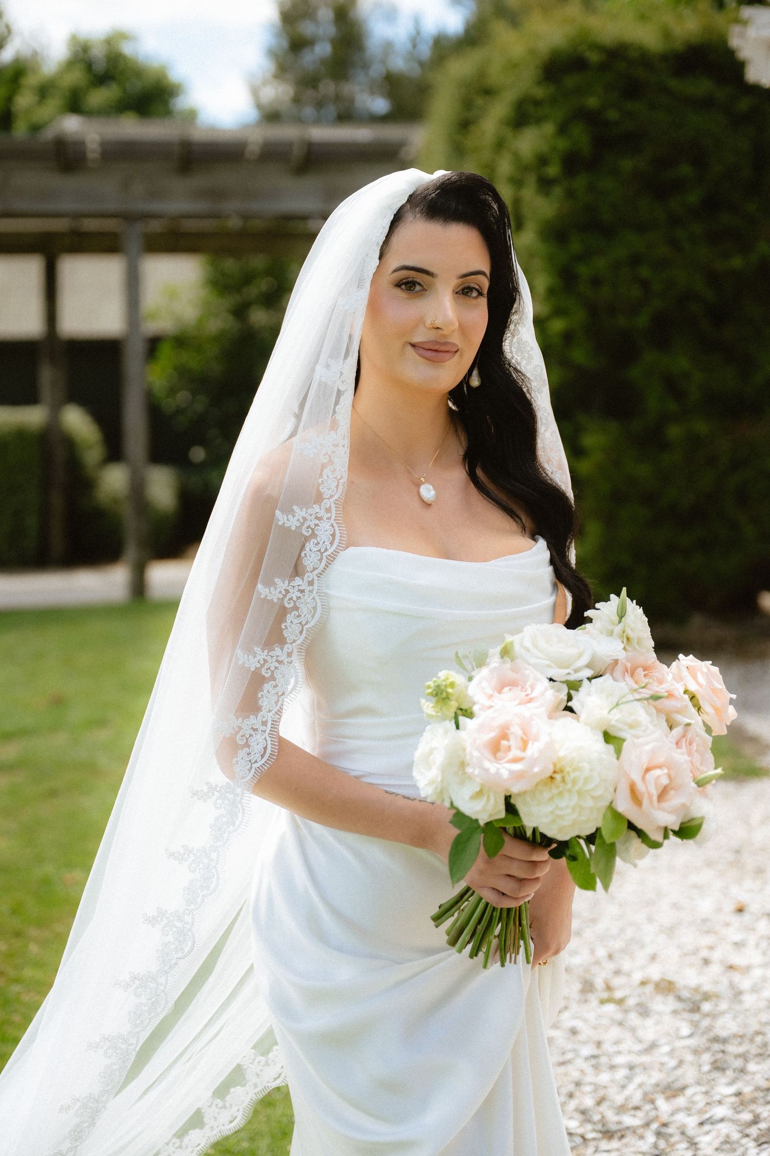 Elegant bride in white strapless gown and lace veil holding blush pink and white rose bouquet in garden setting.