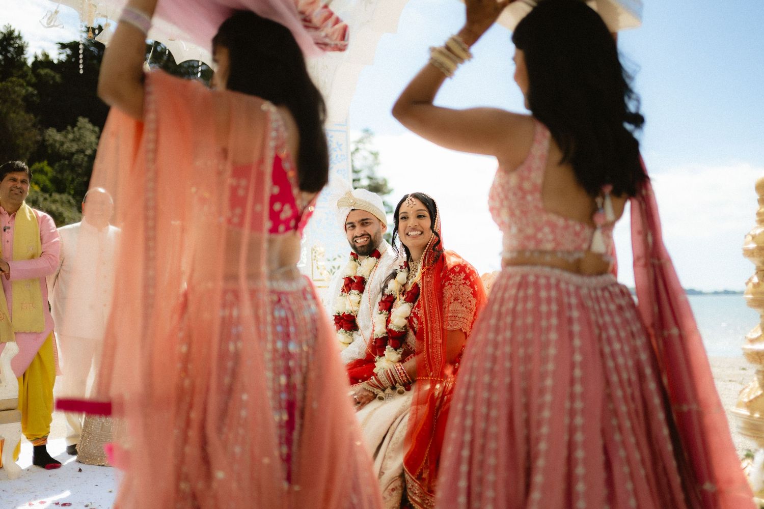 Indian wedding ceremony with bride and groom seated as bridesmaids in pink lehengas hold a floral canopy outdoors.