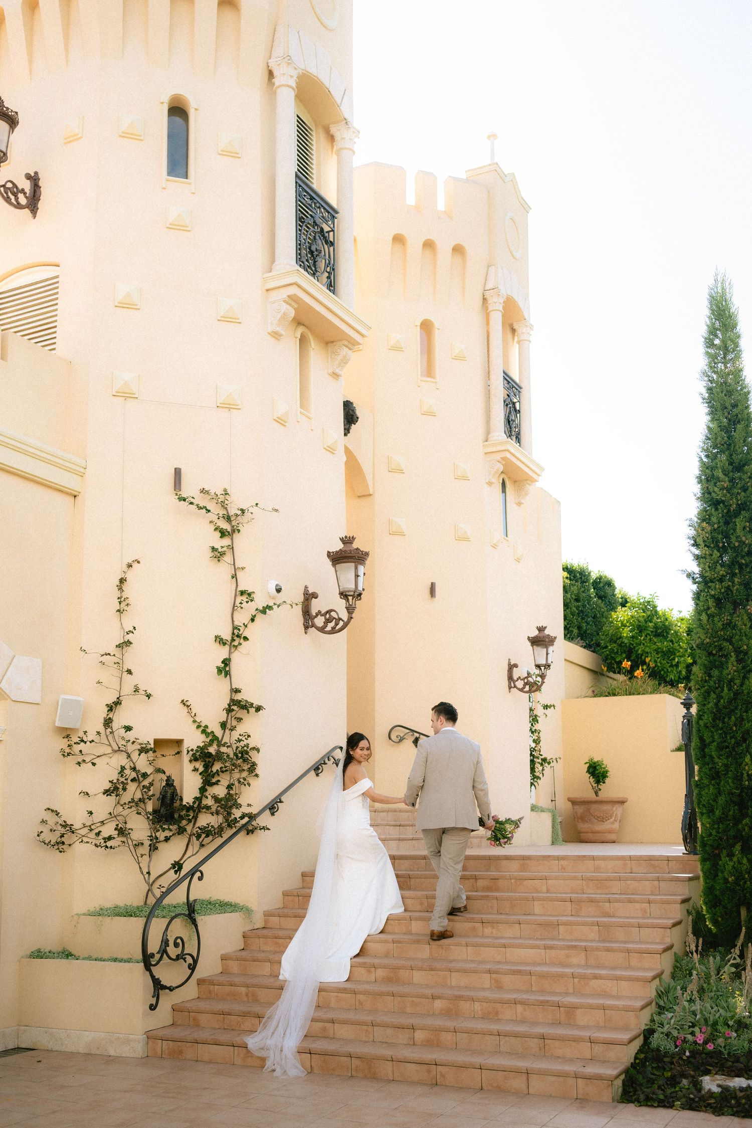 Bride and groom ascending stone steps of an elegant cream castle venue, surrounded by cypress trees.
