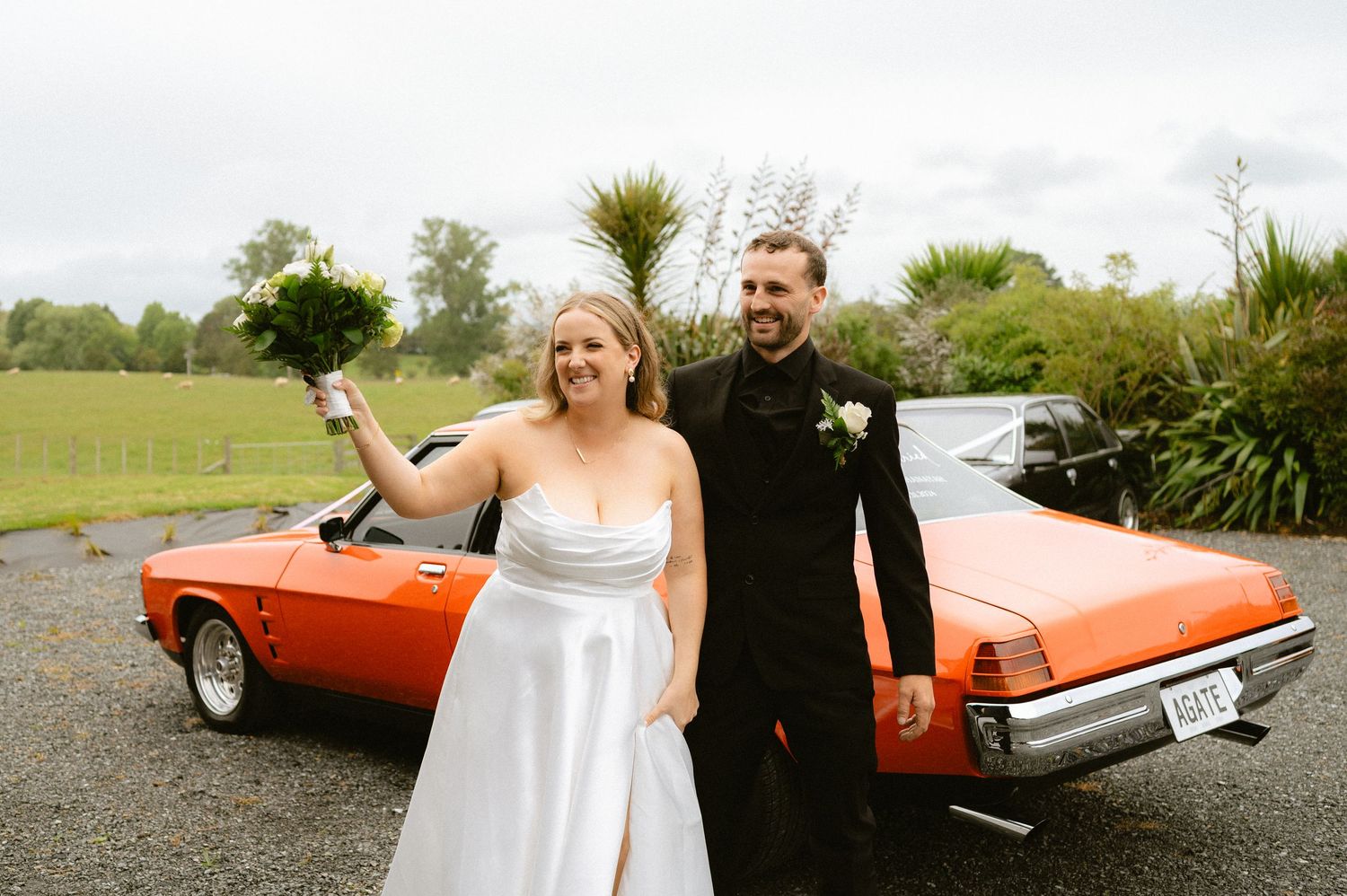 Wedding couple posing with a bright orange vintage convertible car in a countryside setting.