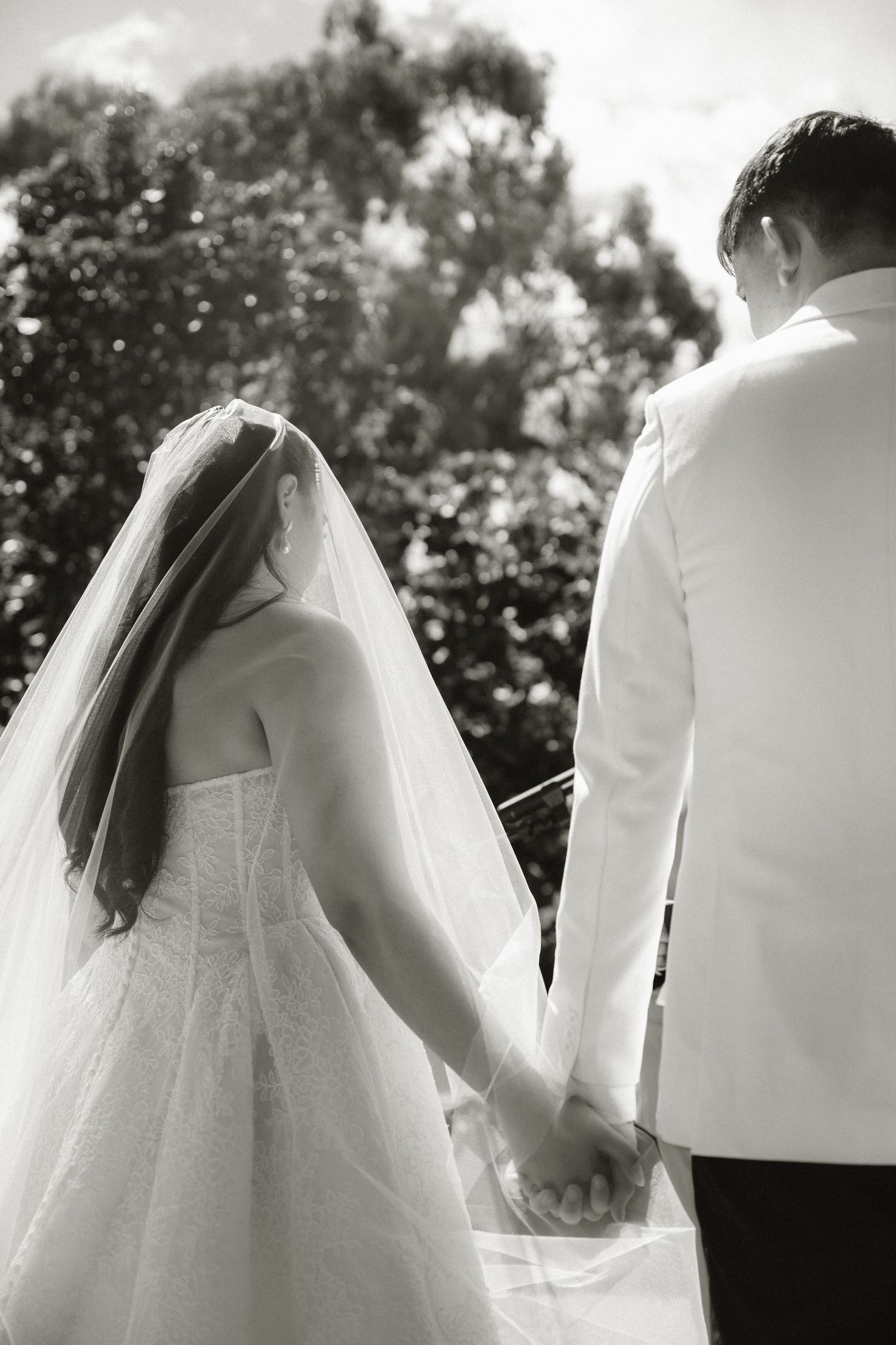 Black and white photo of bride and groom holding hands, walking outdoors, bride in white gown with veil.