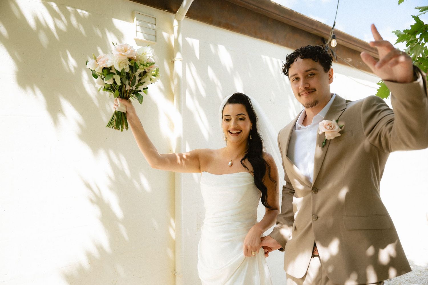 Happy newlyweds celebrate outdoors, bride in white strapless gown holding floral bouquet, groom in tan suit making peace sign.