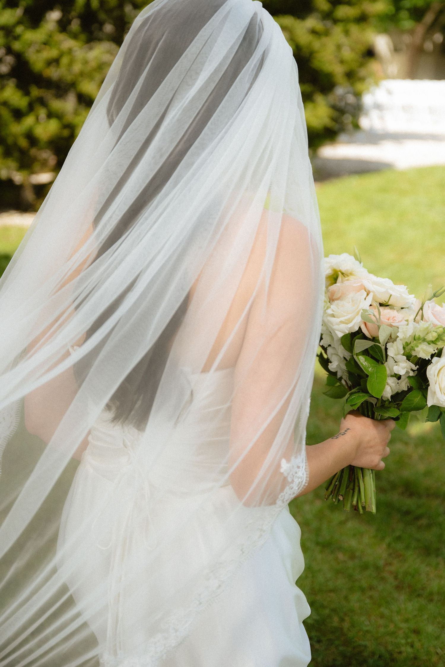 Bride in white wedding dress and flowing veil holding elegant bouquet of white and blush roses outdoors.