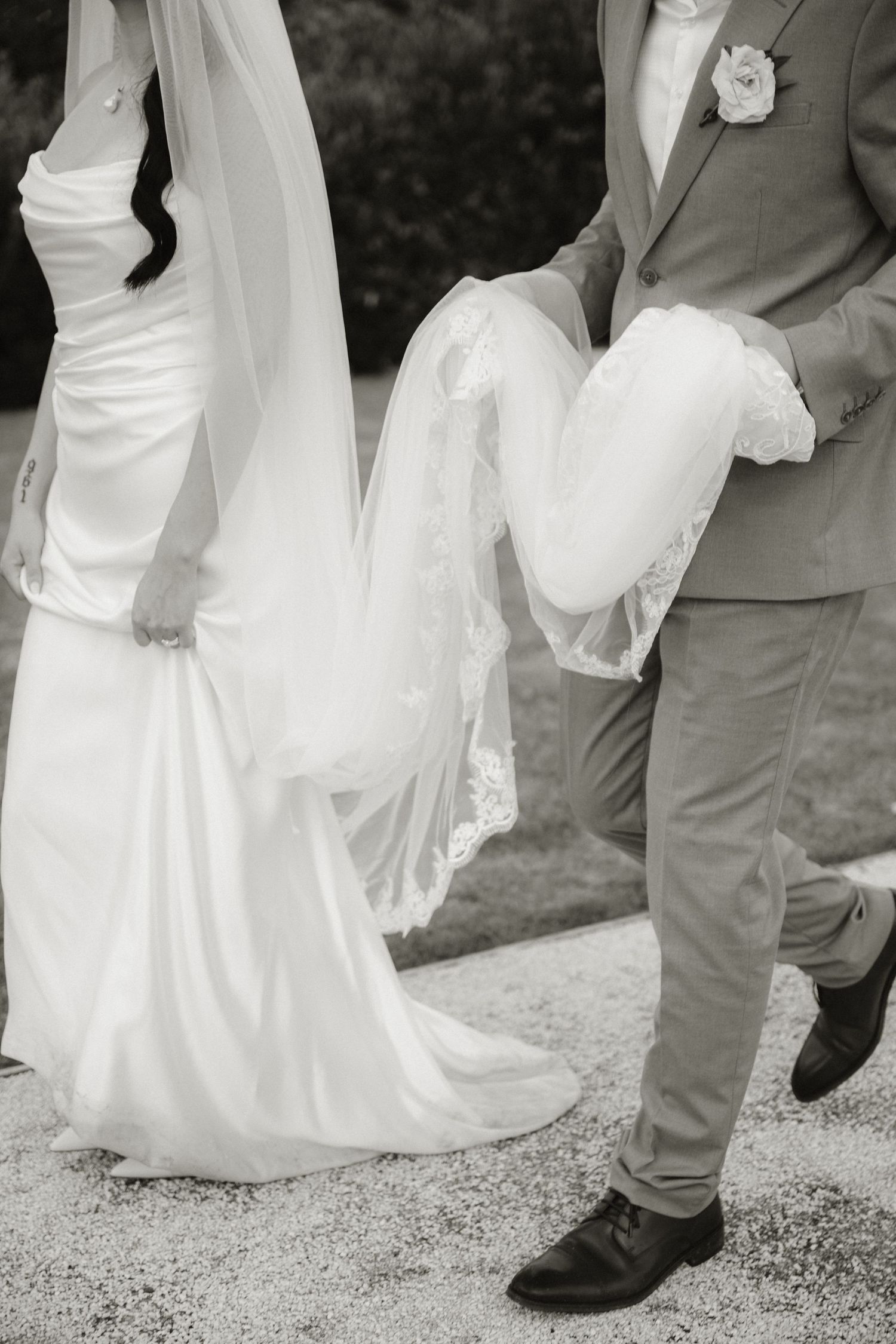 Black and white photo of bride in white gown with groom holding her veil train at outdoor wedding.