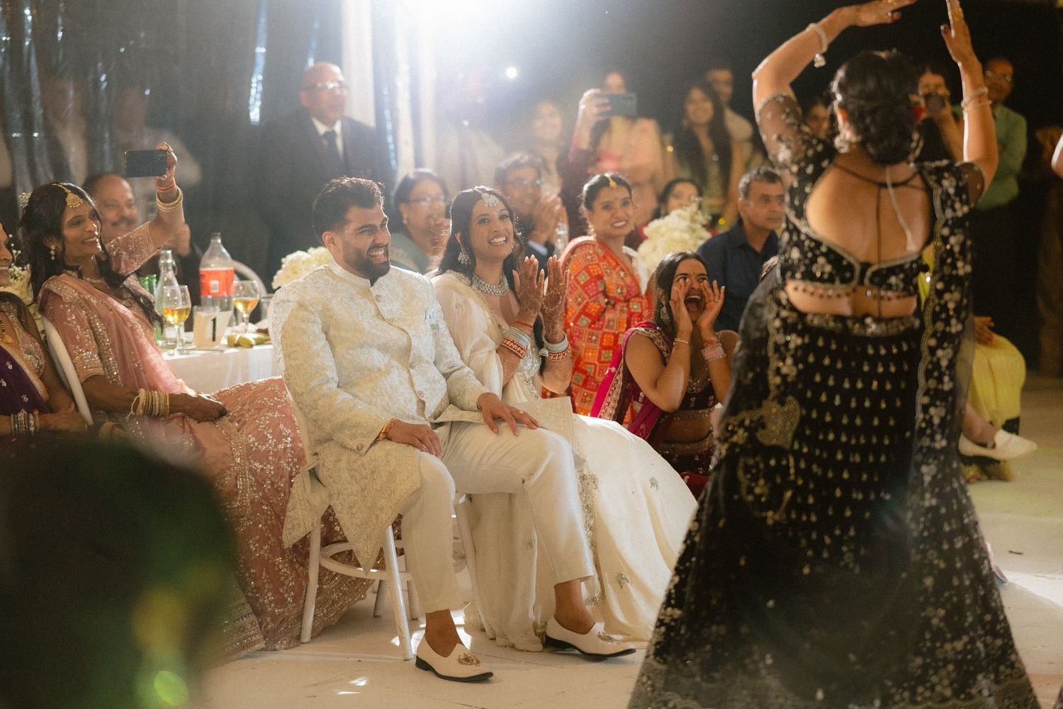 Guests clap and watch a woman performing a dance at an Indian wedding celebration indoors.