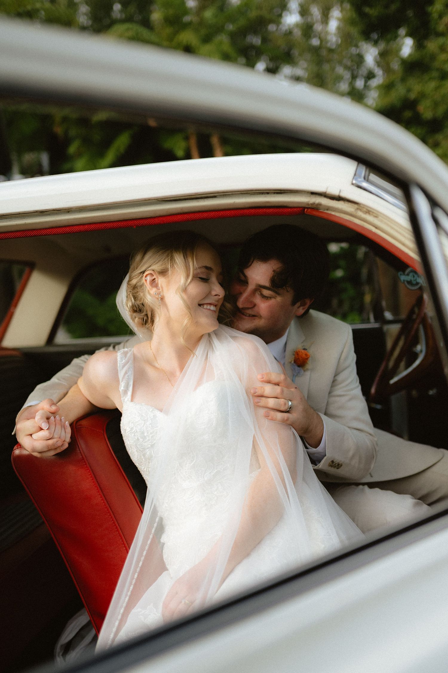 Bride and groom share a romantic moment in a vintage car with red seats, surrounded by lush greenery on their wedding day.