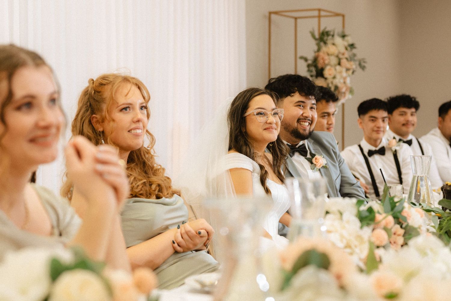 Bride and groom sitting alongside their bridal party, smiling at the person giving a speech.