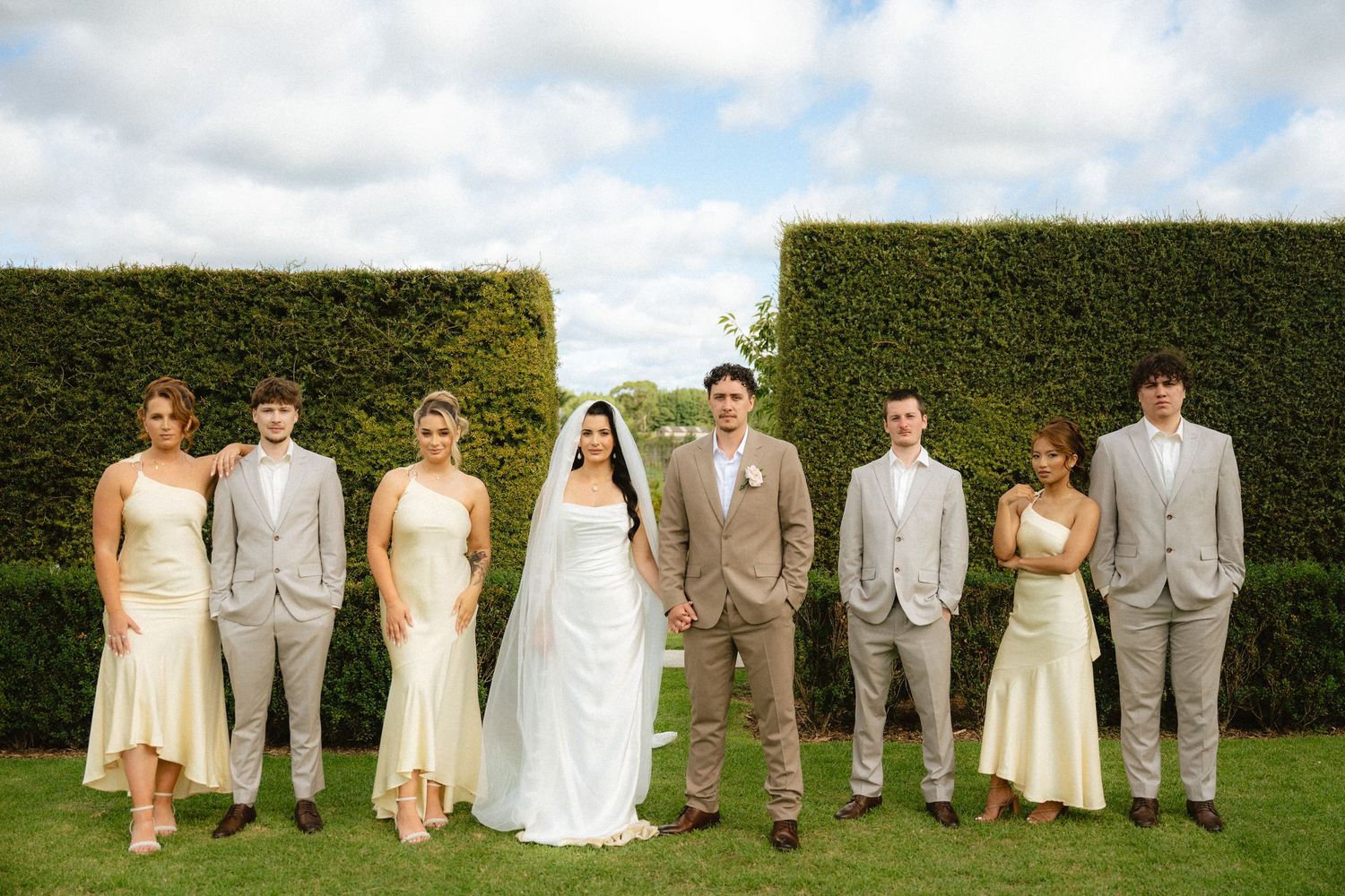 Wedding party poses on green lawn before tall hedge backdrop, bride in white gown flanked by bridesmaids and groomsmen.