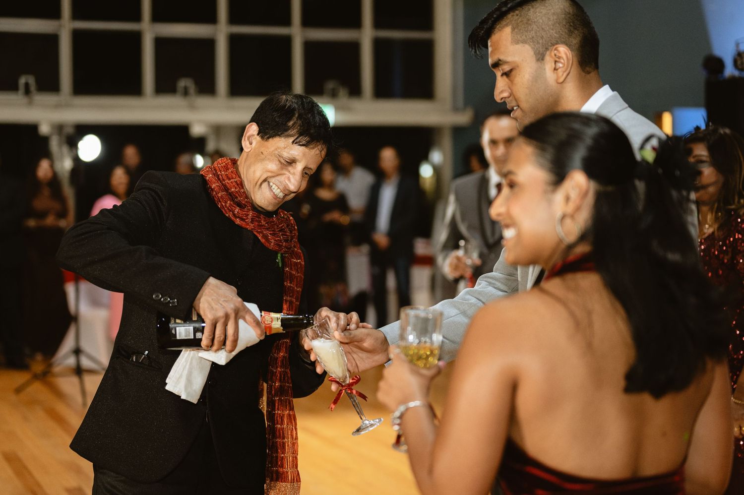 Man with a red scarf pouring champagne into a glass with a red ribbon.