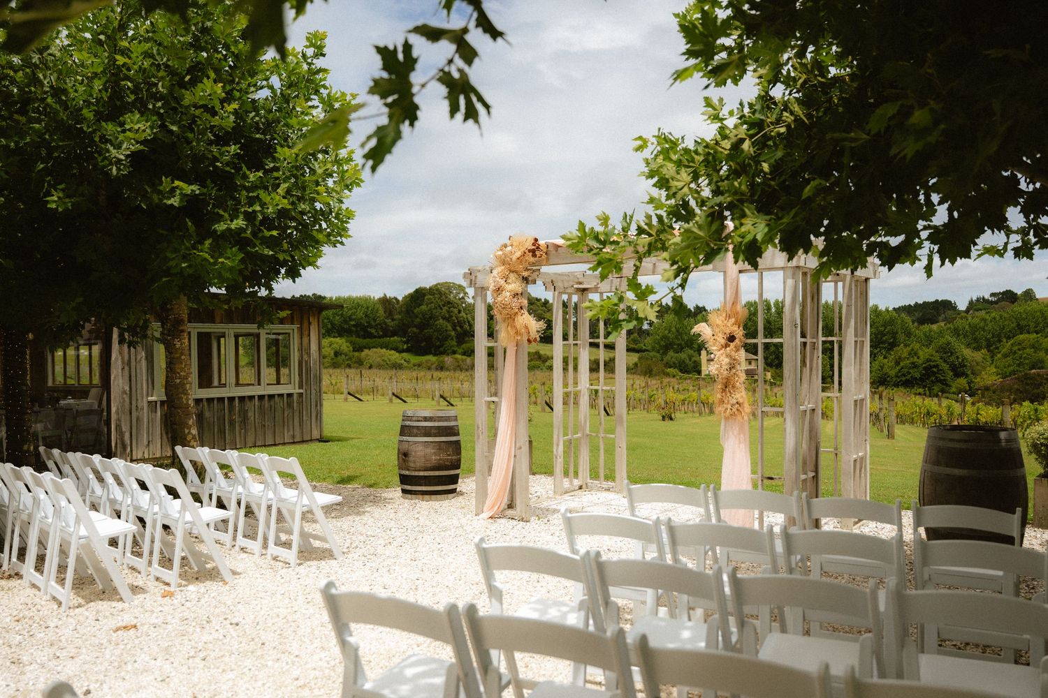 Outdoor wedding ceremony setup with white chairs, wooden barrels, and floral-decorated arbor surrounded by lush green trees.