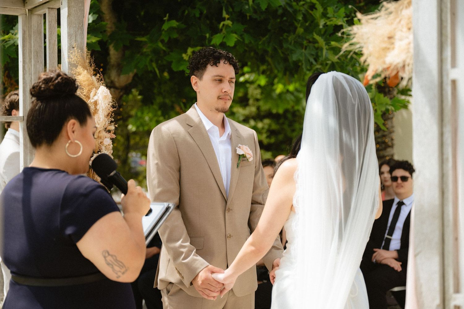 Bride and groom hold hands during outdoor wedding ceremony while officiant reads vows to smiling guests.