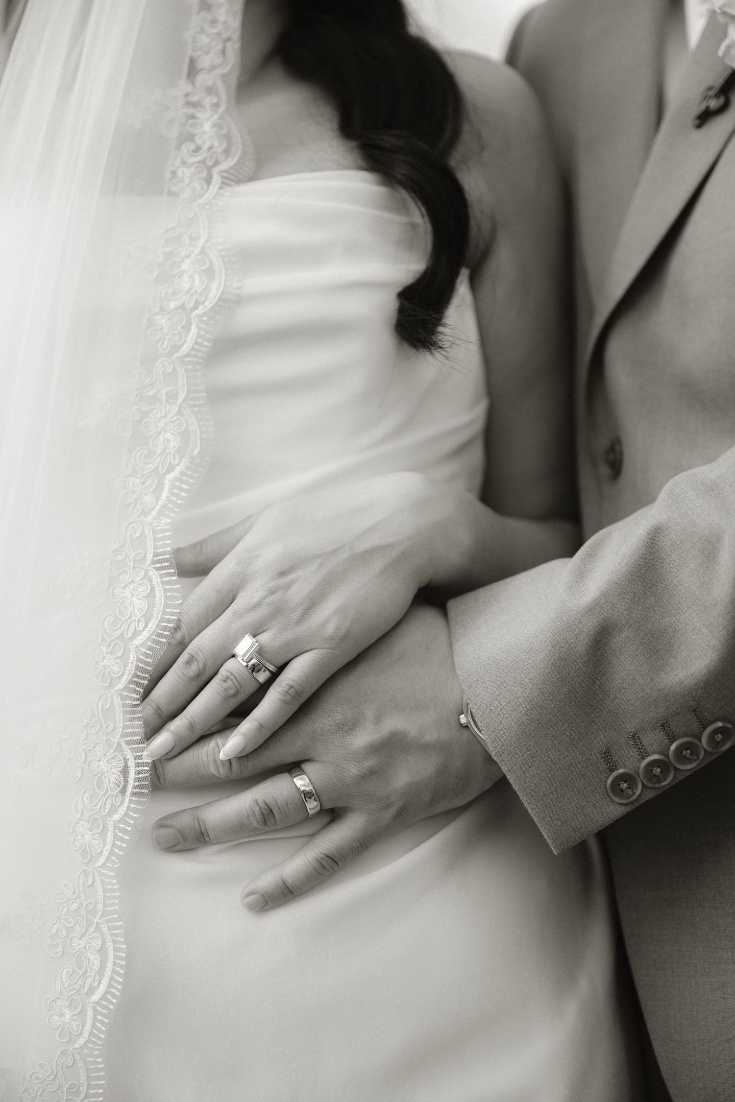 Black and white wedding photo of bride and groom hands showing rings, with lace veil detail.