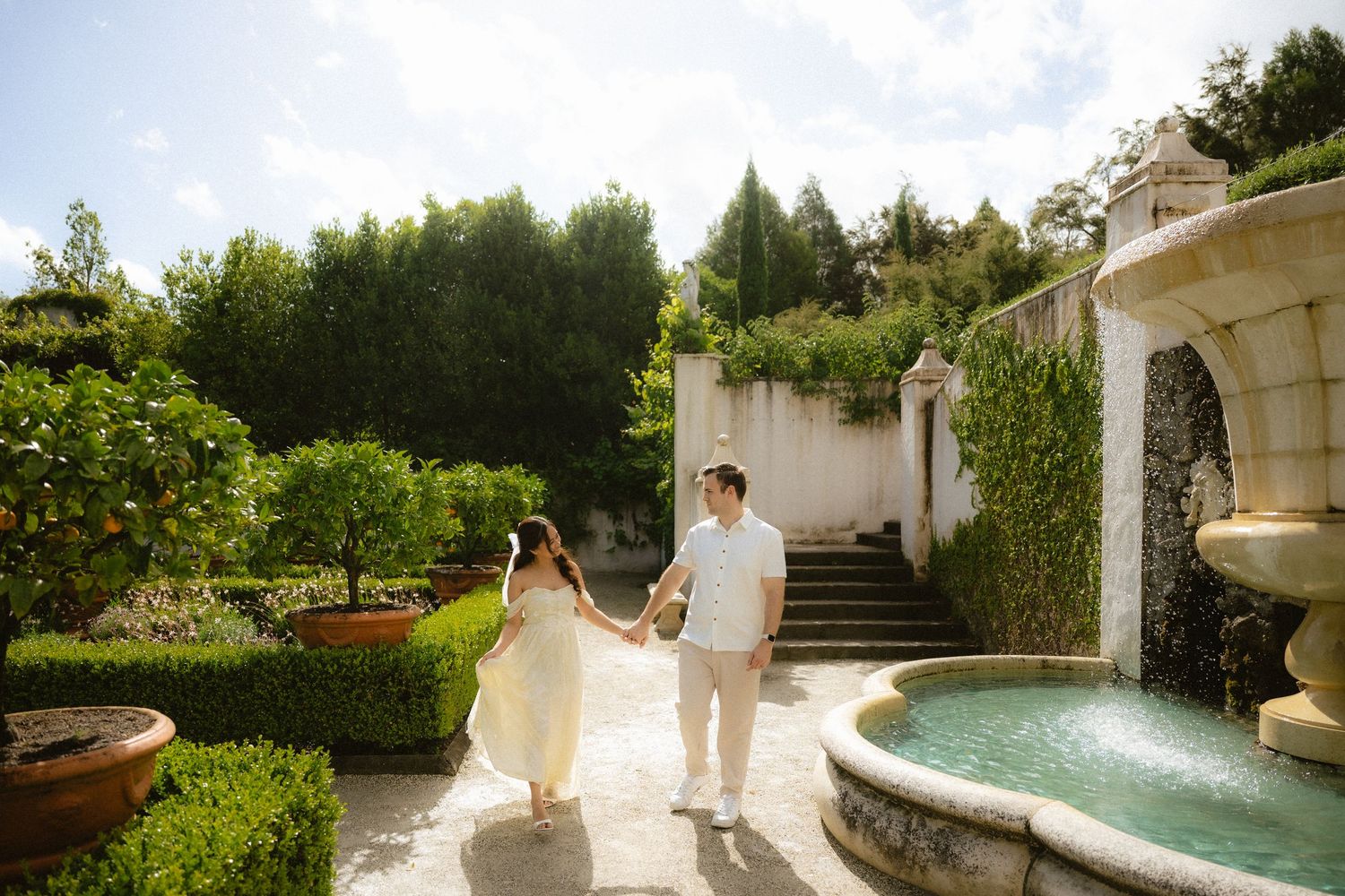 Couple holding hands walking through an elegant garden with a fountain, lush greenery, and stone walls on a sunny day.