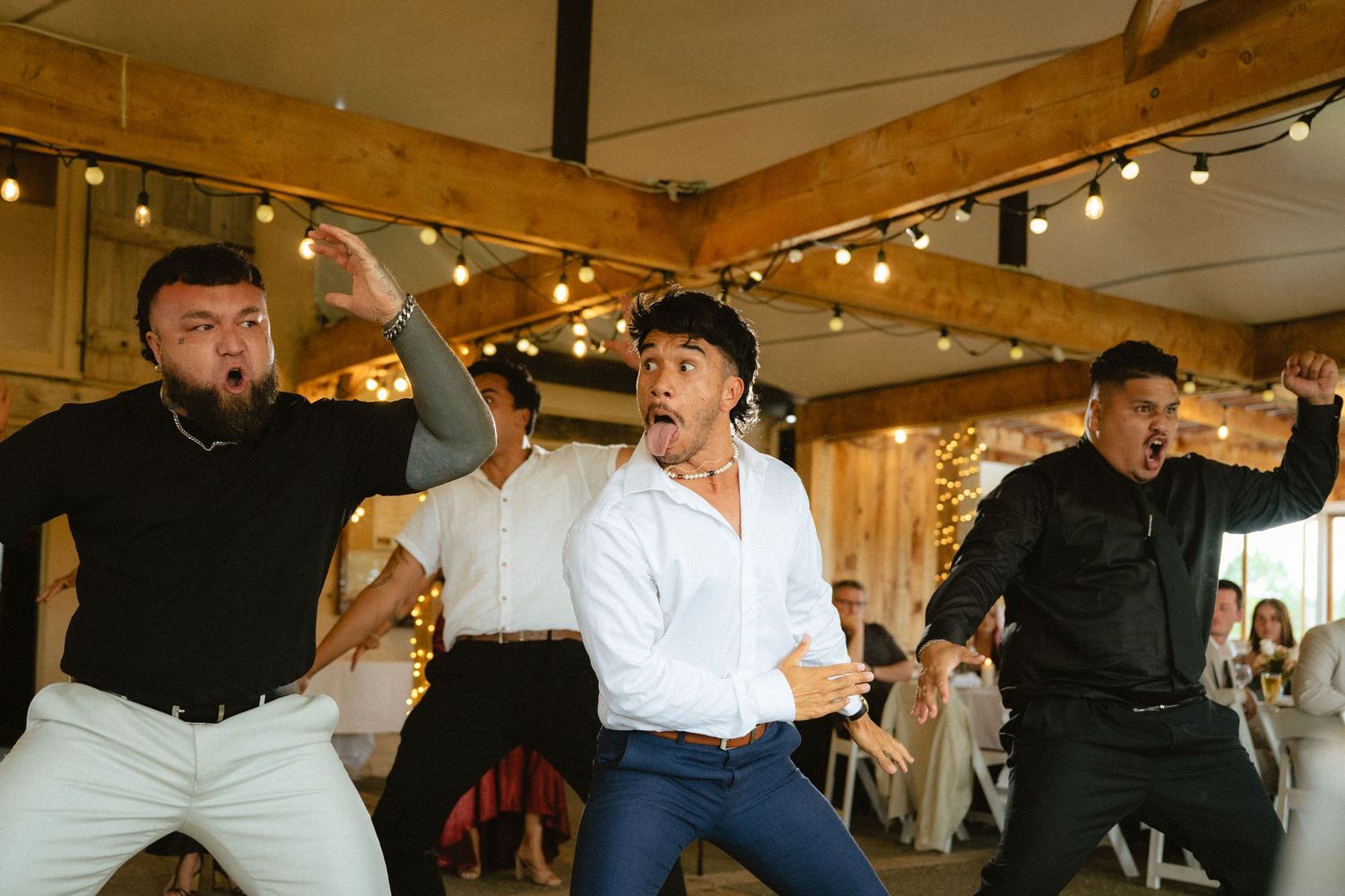 Three men performing a haka dance at a wedding reception in a rustic barn venue with string lights.