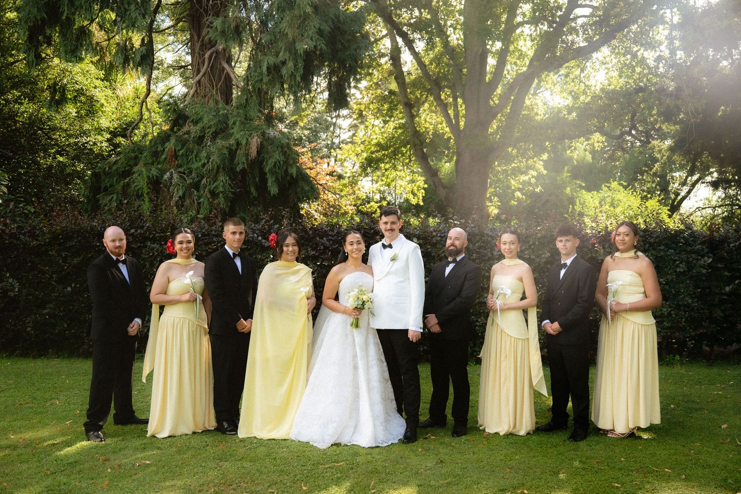 Wedding party poses outdoors in lush garden, bride in white gown with groom and bridesmaids in yellow dresses.