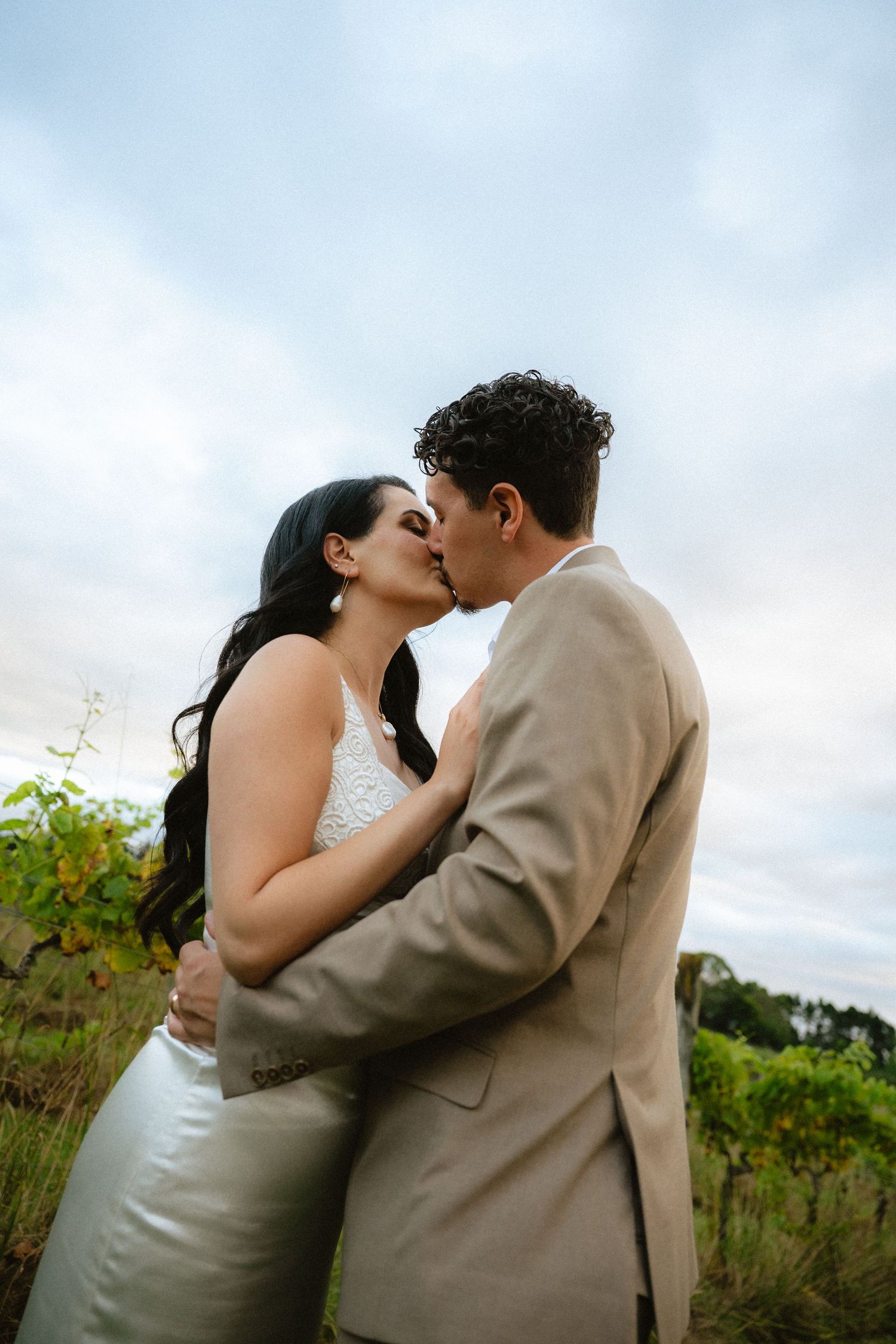 A bride and groom share a romantic kiss outdoors in a vineyard setting under a cloudy sky.