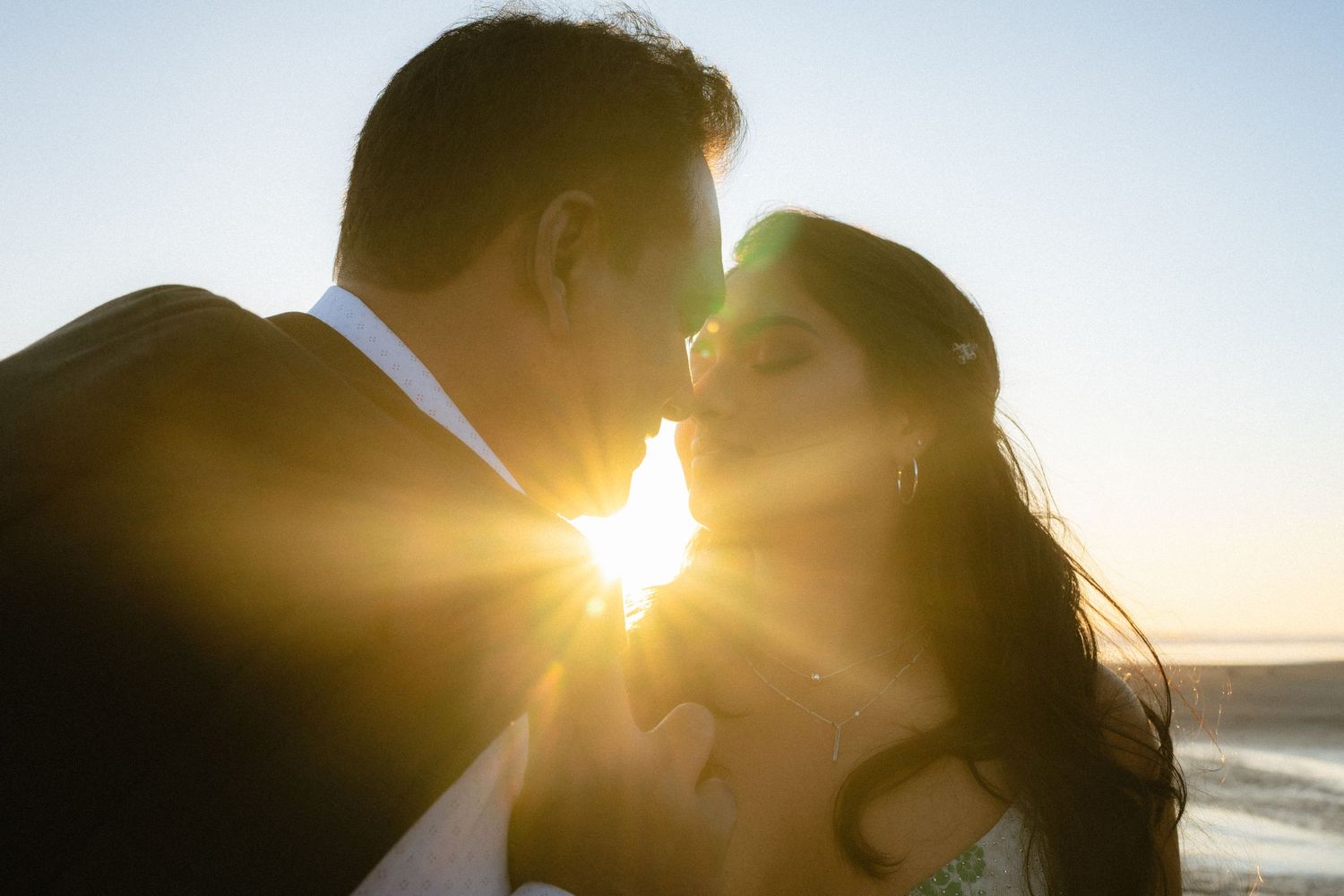 Couple sharing a romantic kiss at sunset on a beach, with golden sunlight glowing between them.