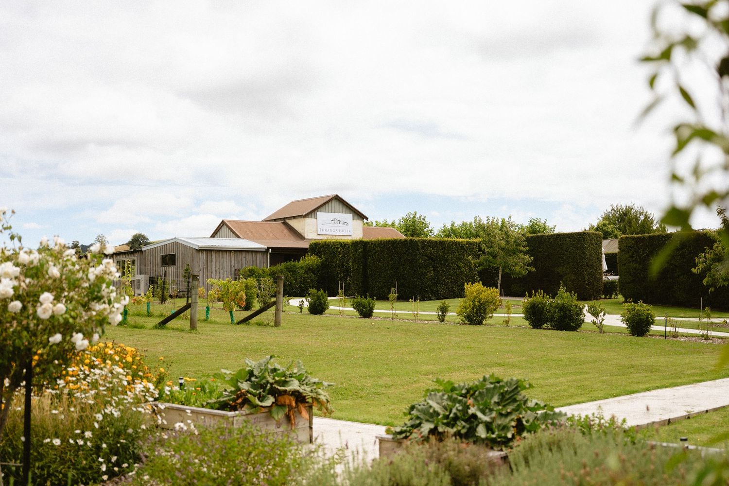 Lush green garden with raised vegetable beds, manicured hedges, and a charming barn-style house under cloudy skies.