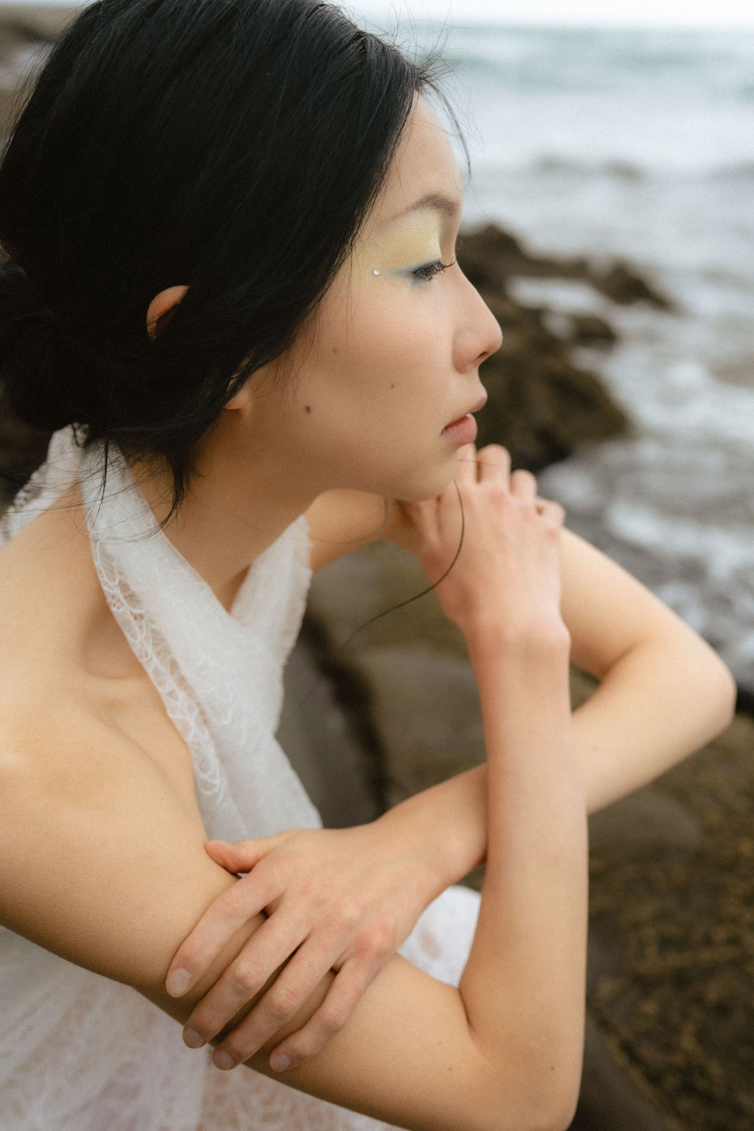 A young Asian woman in a white dress poses thoughtfully by rocky ocean shores, wearing artistic eye makeup.