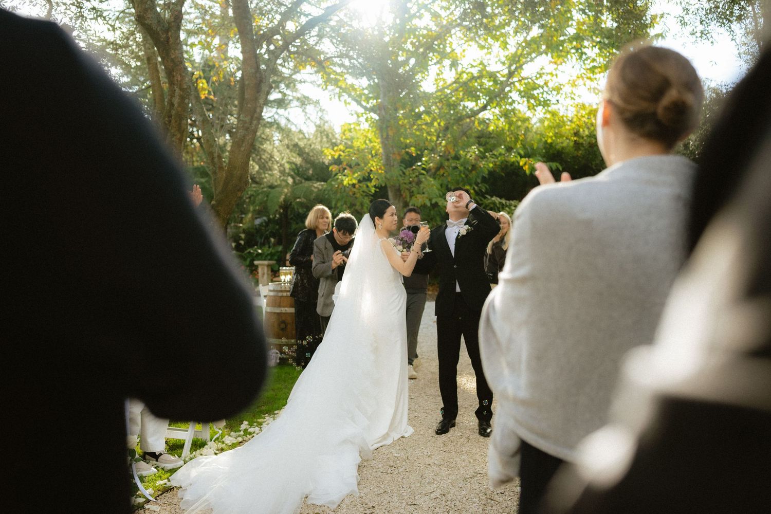Bride and groom share first kiss at outdoor wedding ceremony surrounded by guests and lush green trees.