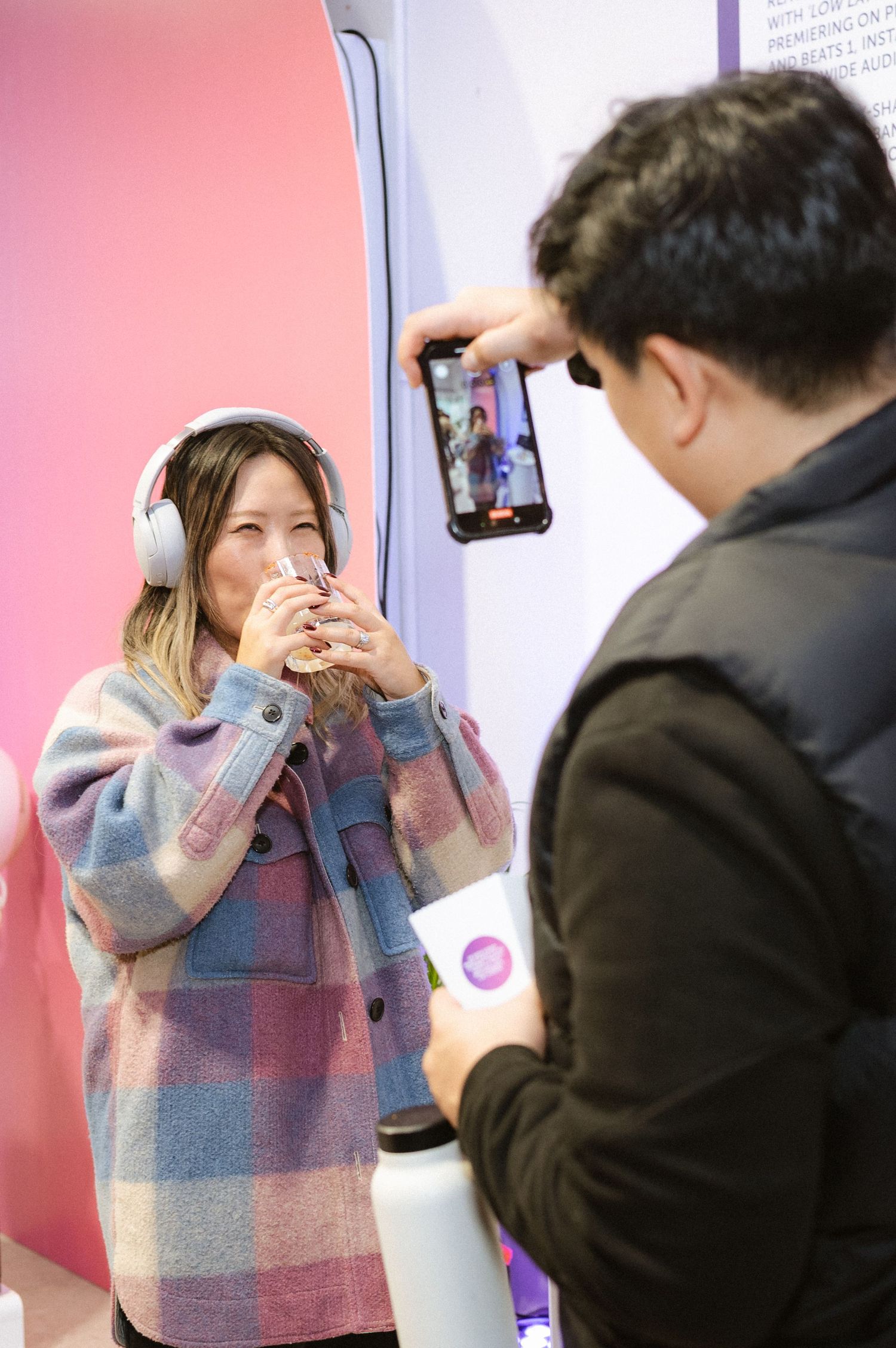 A man photographs a smiling woman wearing headphones and a plaid coat sipping a drink at a colorful pink event.