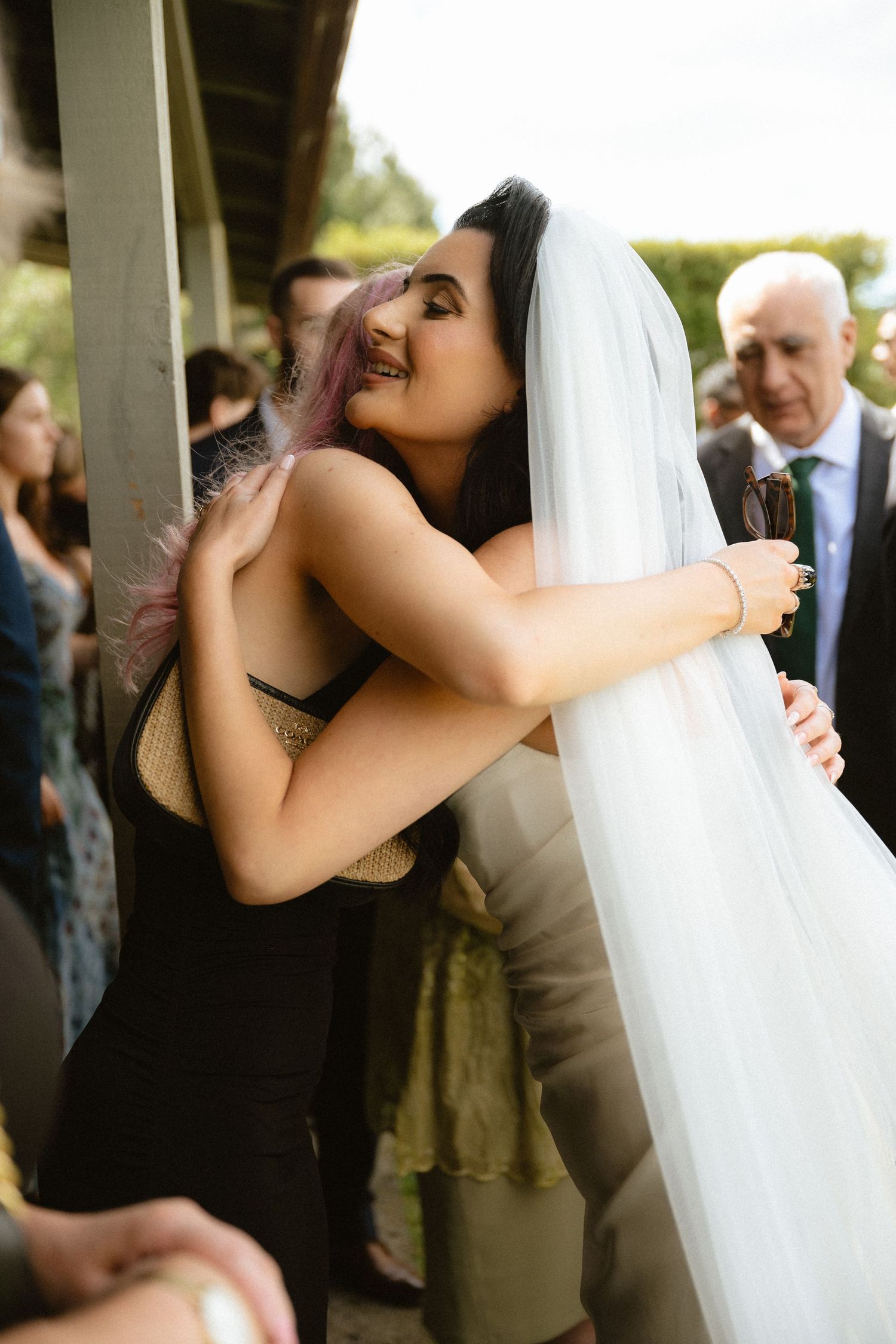 A bride wearing a veil hugs a guest with purple hair at an outdoor wedding reception.