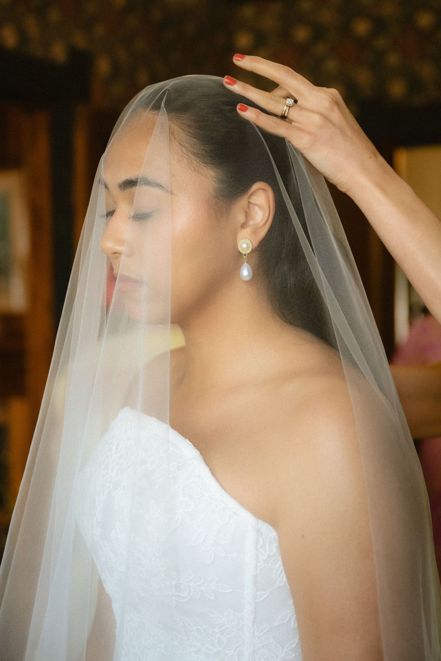 Bride in white strapless gown with sheer veil being placed, wearing pearl drop earrings, eyes closed, serene expression.