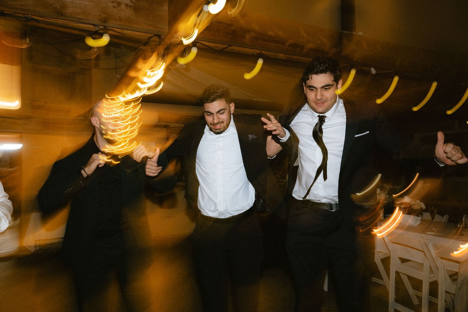 Three men in suits dancing joyfully at a wedding reception with warm bokeh lights in the background.