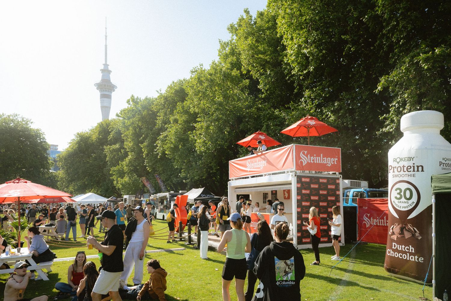 Outdoor event setup with white tents and red banners along a tree-lined path with the Sky Tower visible in background.