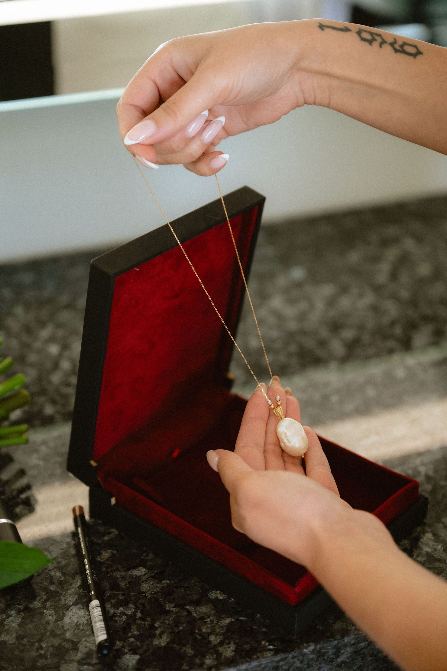 Hands holding a gold oval pendant necklace over a red velvet jewelry box on a granite surface.
