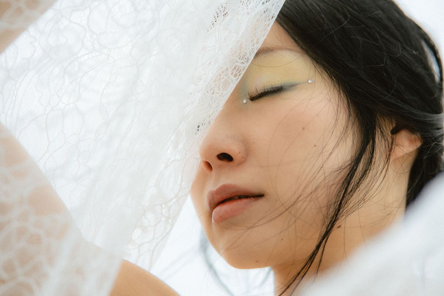 Close-up of an Asian woman with eyes closed, partially covered by a delicate white lace veil.