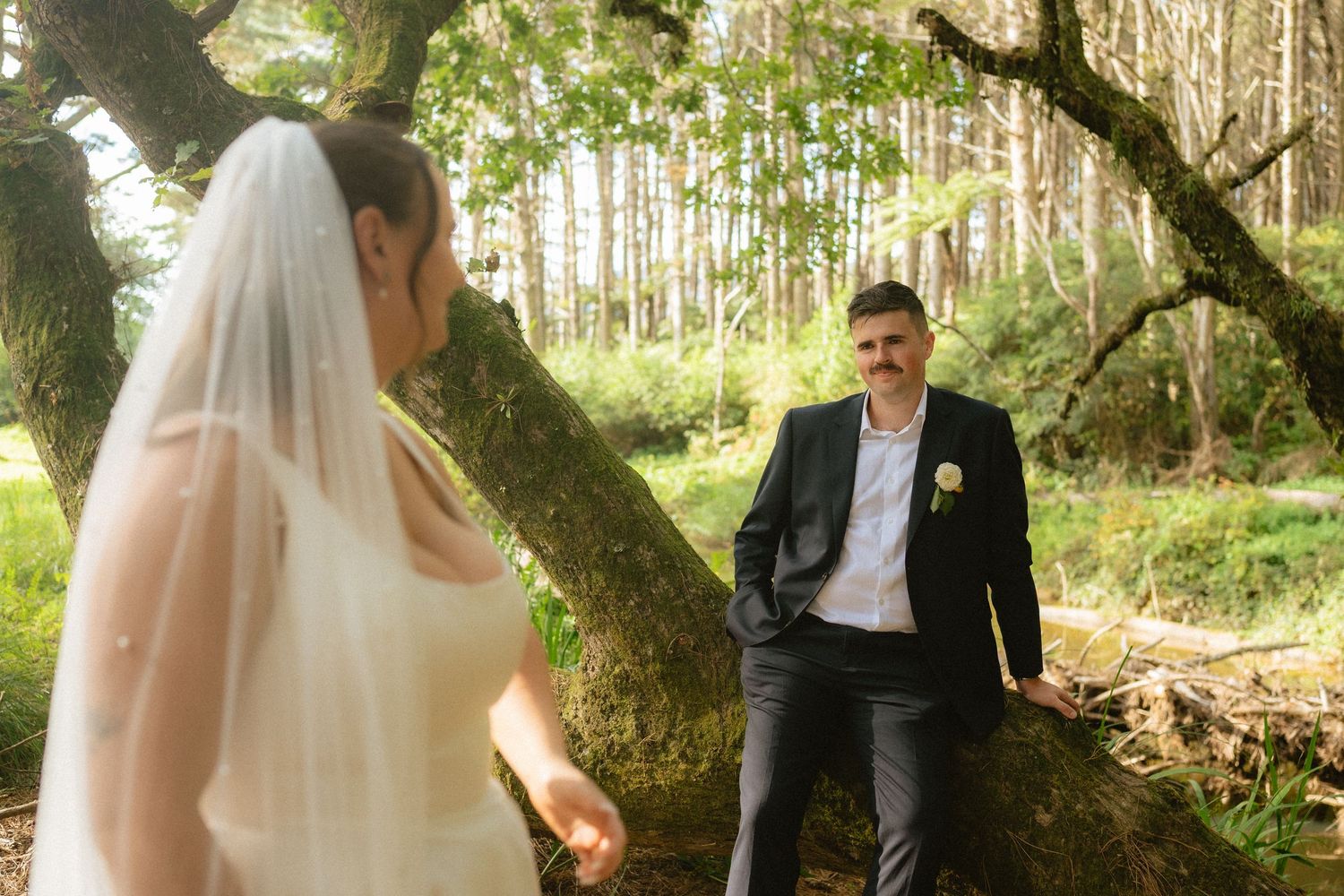 Bride in white gown and veil stands near groom in dark suit leaning on fallen tree in lush forest wedding photo.