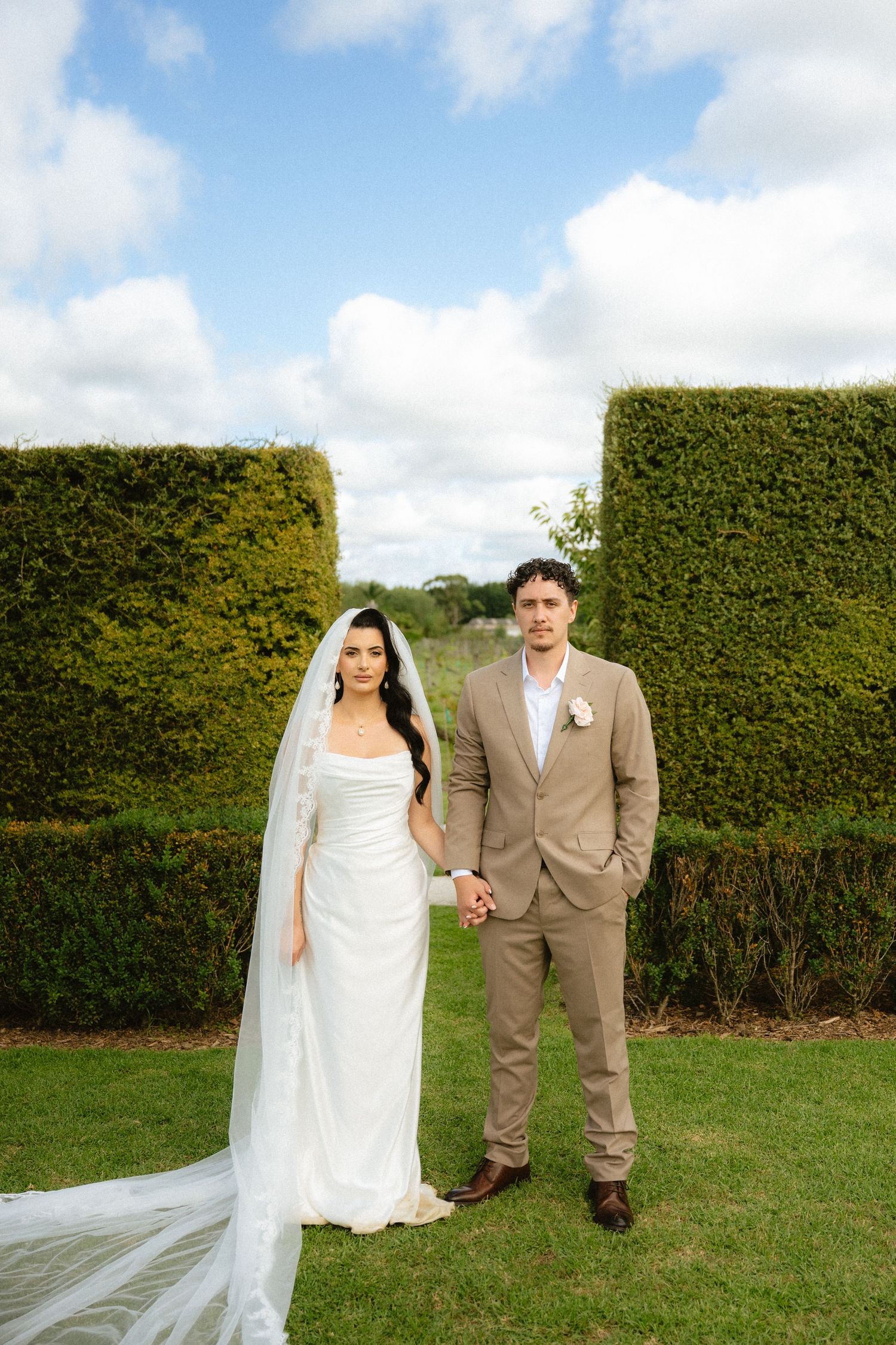 Bride in white gown with veil and groom in tan suit hold hands in a garden with tall hedges.