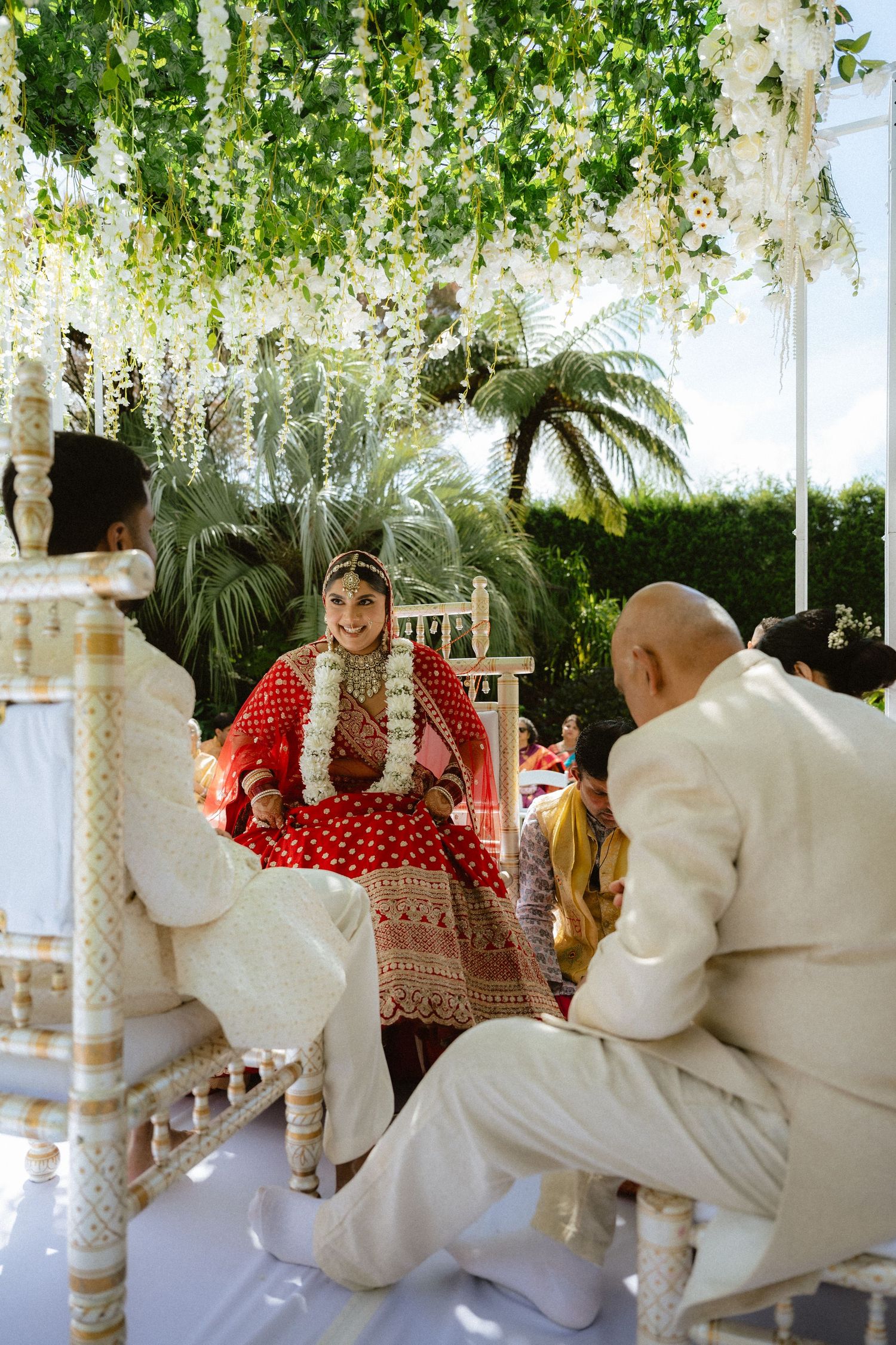 Smiling Indian bride in red lehenga with floral garland at outdoor wedding ceremony under white flower canopy.