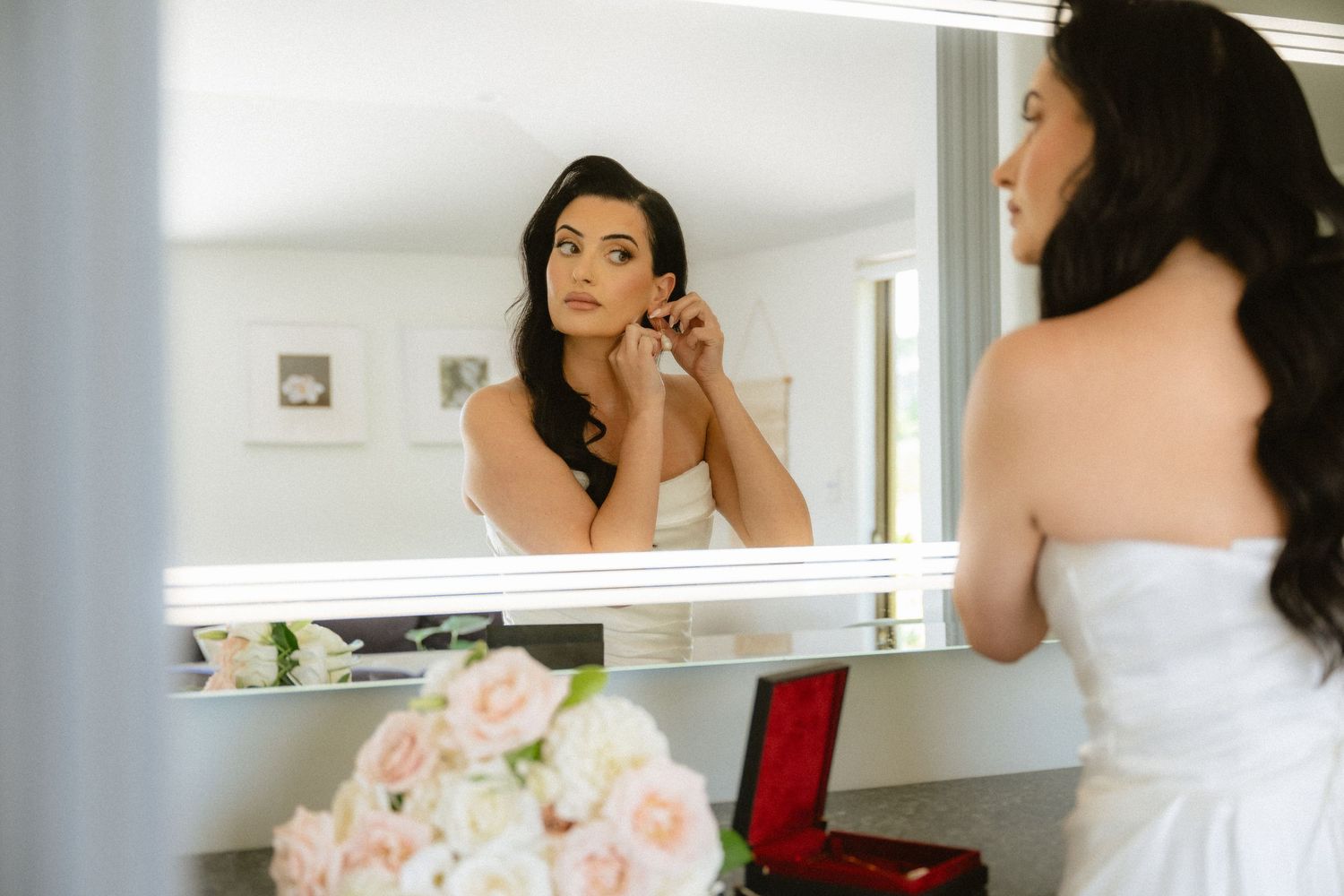 Bride in strapless white gown applying earrings in a lighted mirror, with pink roses nearby.
