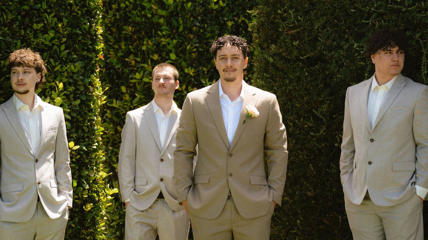 Four groomsmen in matching tan suits posing outdoors in front of a lush green hedge wall at a wedding.