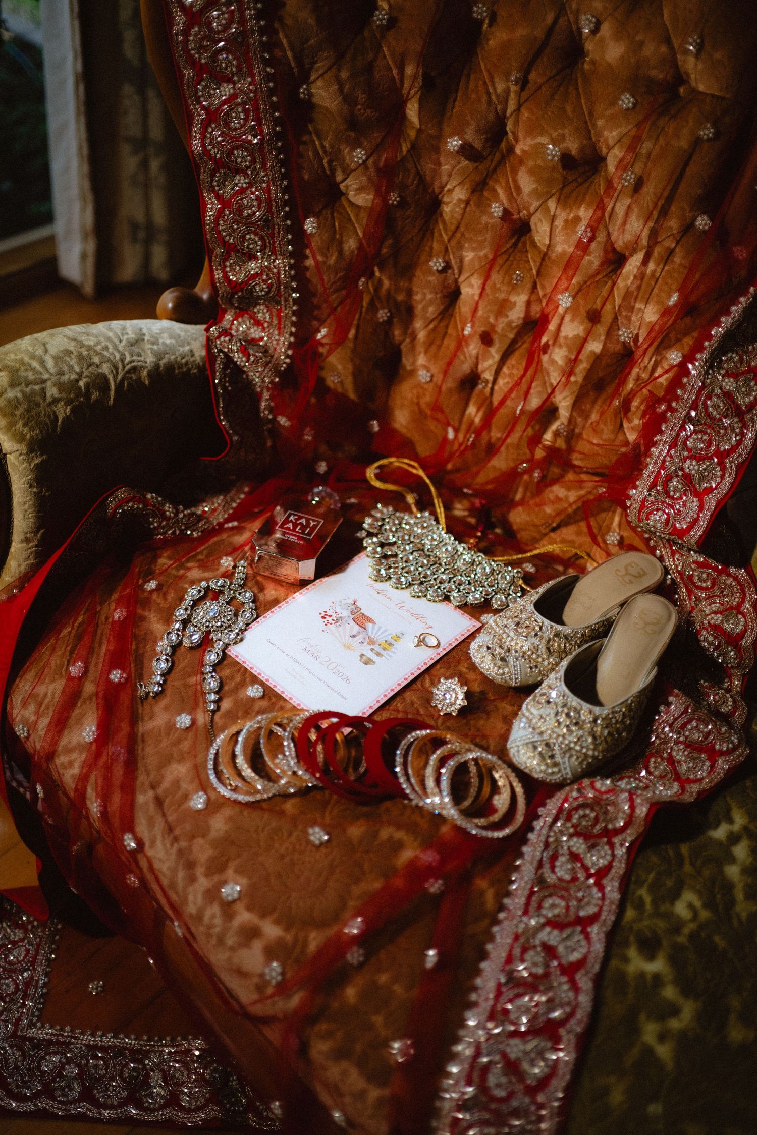 Traditional Indian bridal accessories on ornate chair, including bangles, jewelry, embroidered juttis, and red dupatta.