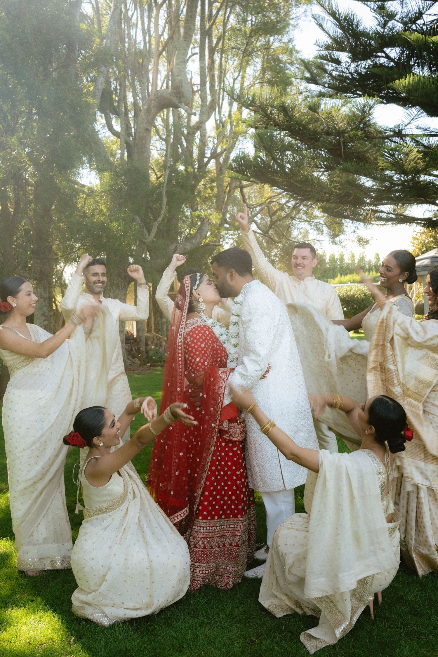 Indian bride in red sari and groom in white kurta celebrated by wedding party in cream sarees outdoors.