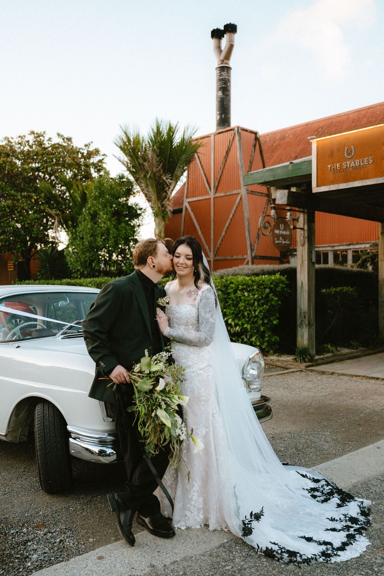 Bride and groom pose by a vintage white car outside The Stables venue, bride holding a lush greenery bouquet.