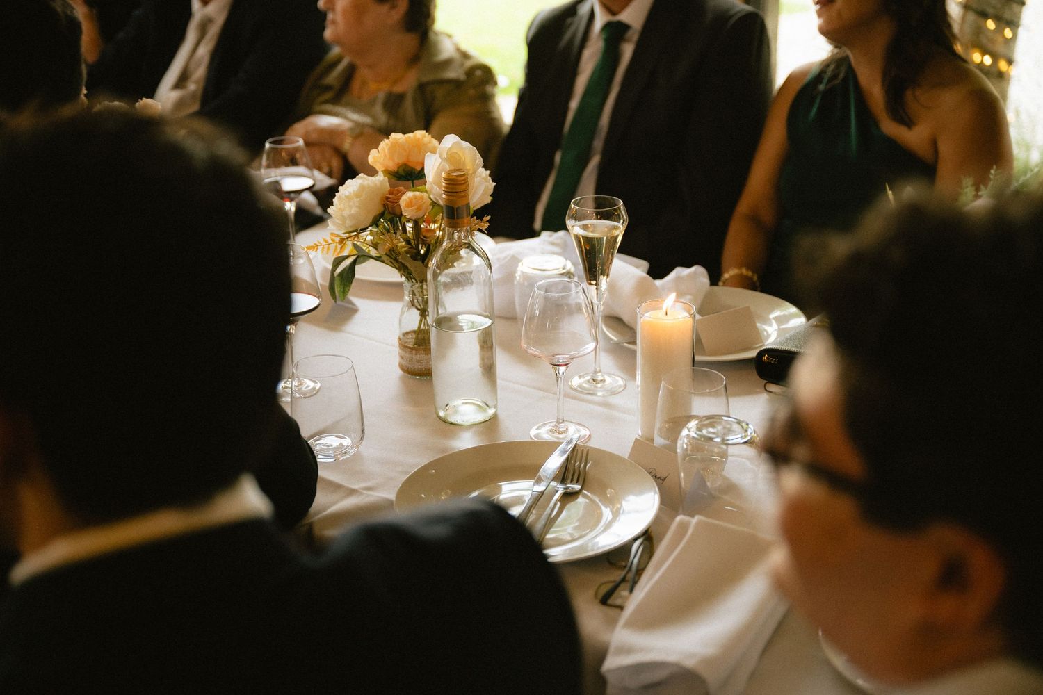 Elegant wedding reception table with white linens, candles, floral centerpiece, and formally dressed guests.