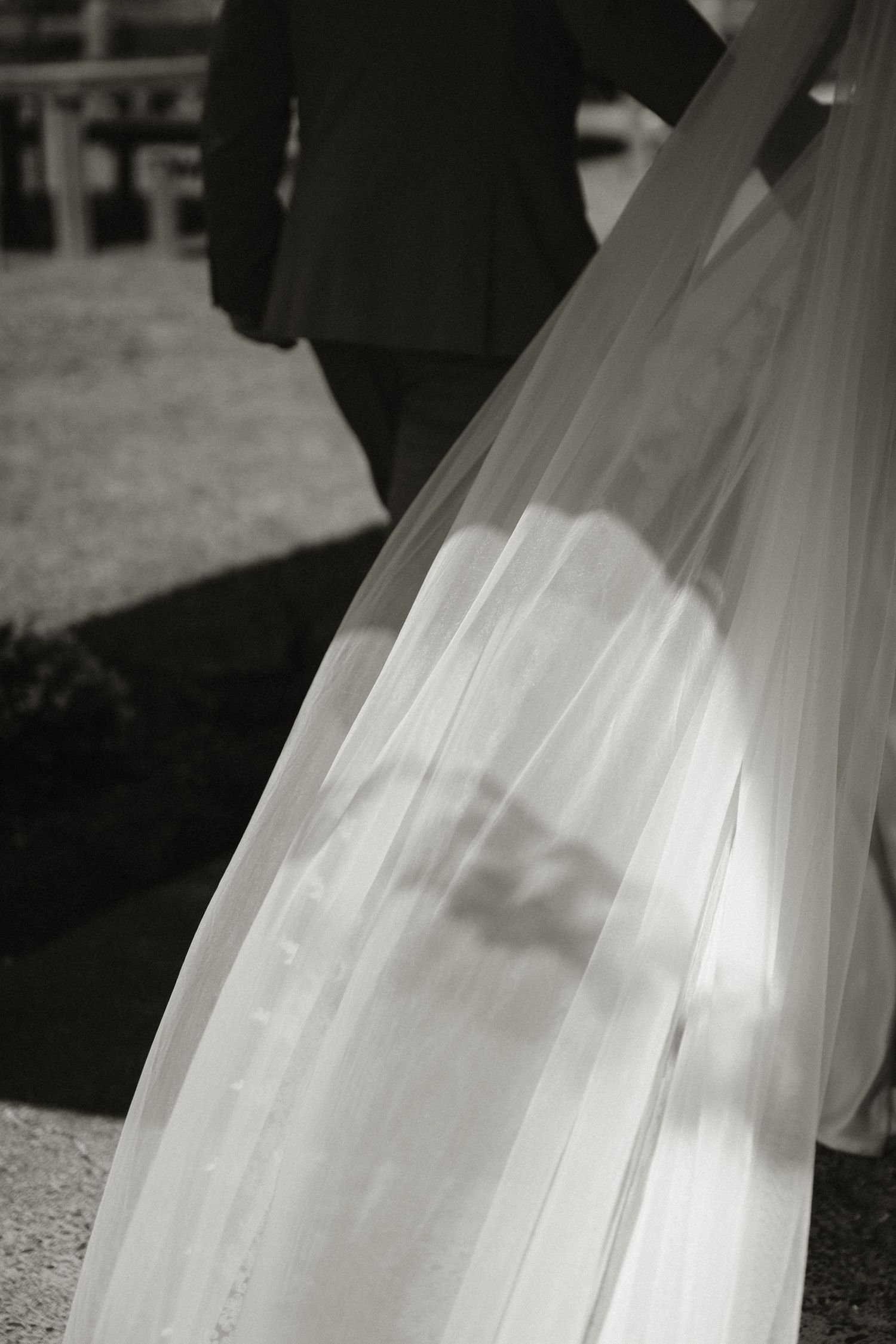 Black and white wedding photo of bride's flowing veil with groom walking ahead on outdoor path.