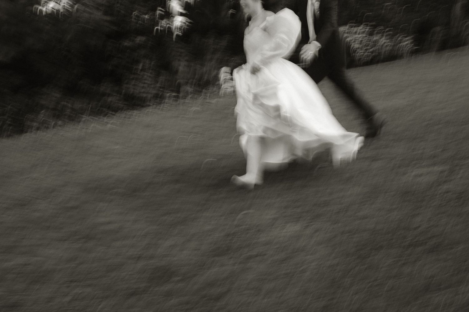 Blurred black and white photo of a bride and groom running across a grassy lawn on their wedding day.