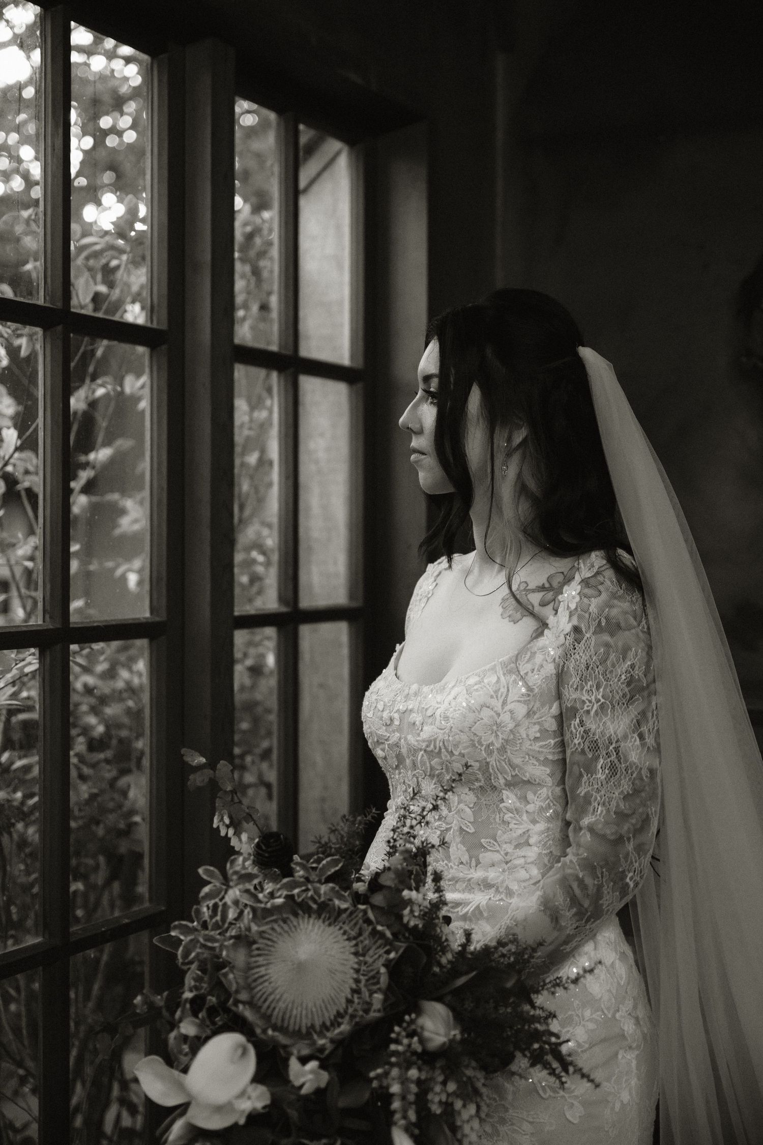 Black and white bridal portrait of a bride in lace gown holding bouquet, gazing out a window with soft natural light.