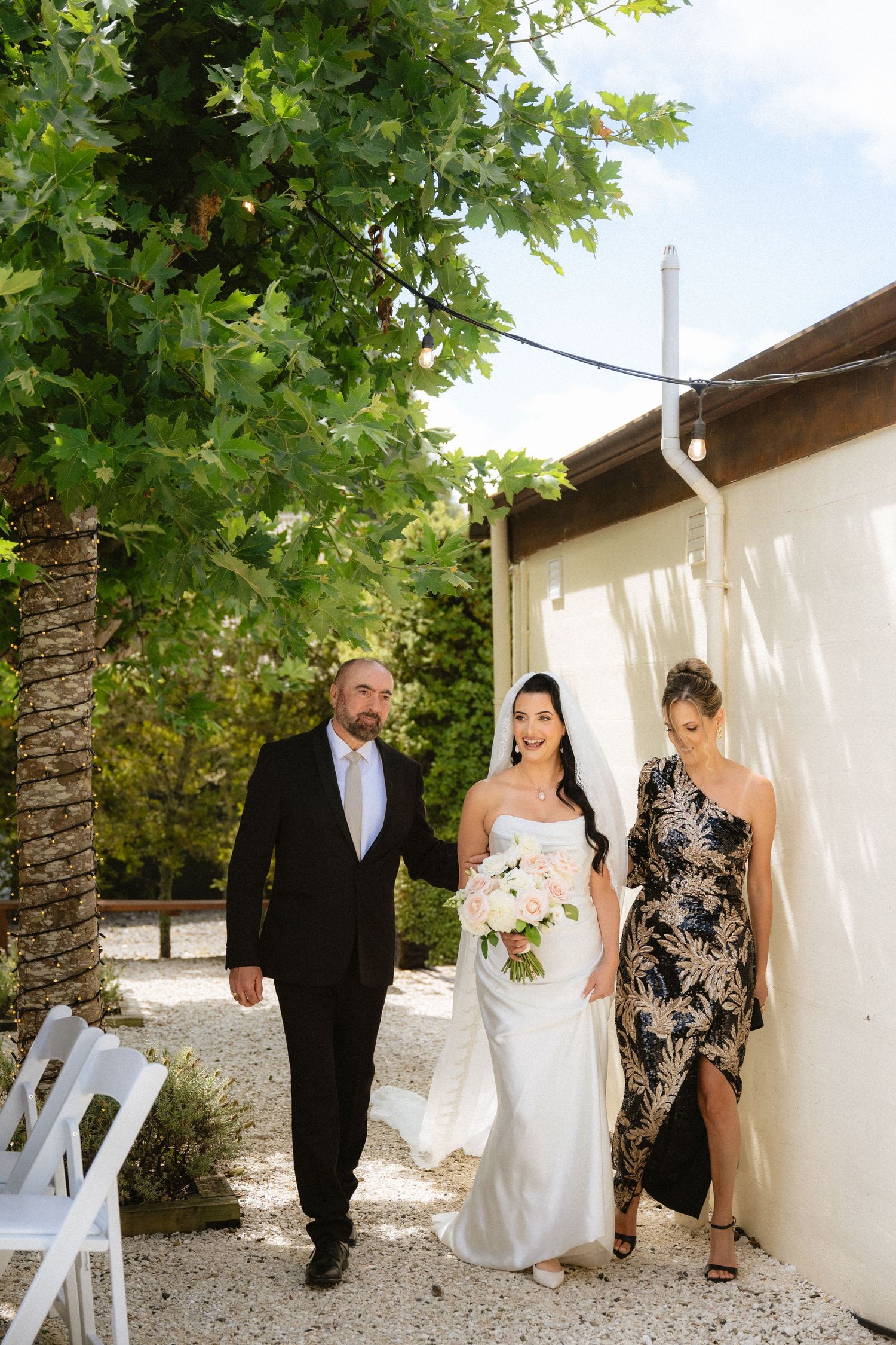 Bride in white gown walks with parents toward outdoor wedding ceremony, holding pink bouquet, flanked by trees.