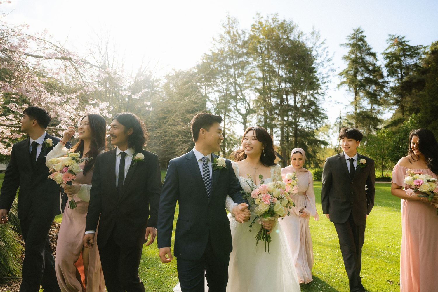 Wedding party stands together outdoors under pink cherry blossoms on a sunny spring day.